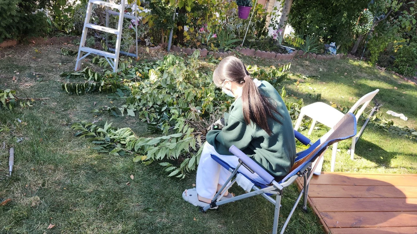 A person sitting on a folding lawn chair in a garden, wearing a face mask and glasses, surrounded by green plants and garden tools.