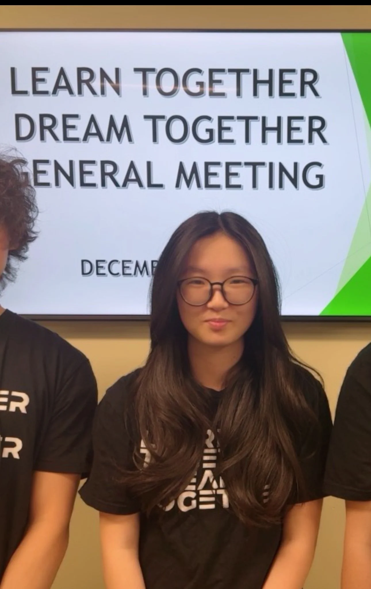 Group of people standing in front of a presentation screen, with a woman in glasses centered, with a sign in the background showing the text 'LEARN TOGETHER DREAM TOGETHER GENERAL MEETING December'.