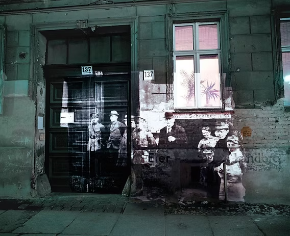 Street art mural on a building wall showing an old black-and-white photo of five people from the early 20th century, with a window and door above.