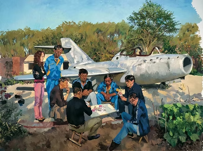 Group of people sitting and standing outdoors near an airplane, playing board games and socializing, with trees and a garden in the background.