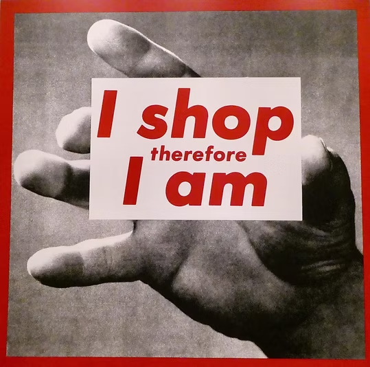 Black and white photo of a hand holding a sign that says "I shop therefore I am" in red and white text.