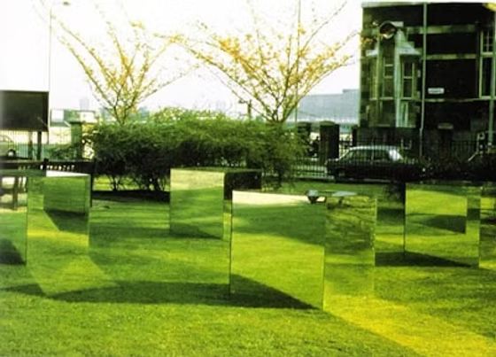 A grassy park area with multiple mirrors placed on the ground reflecting the surrounding trees and sky, with a building and cars in the background.