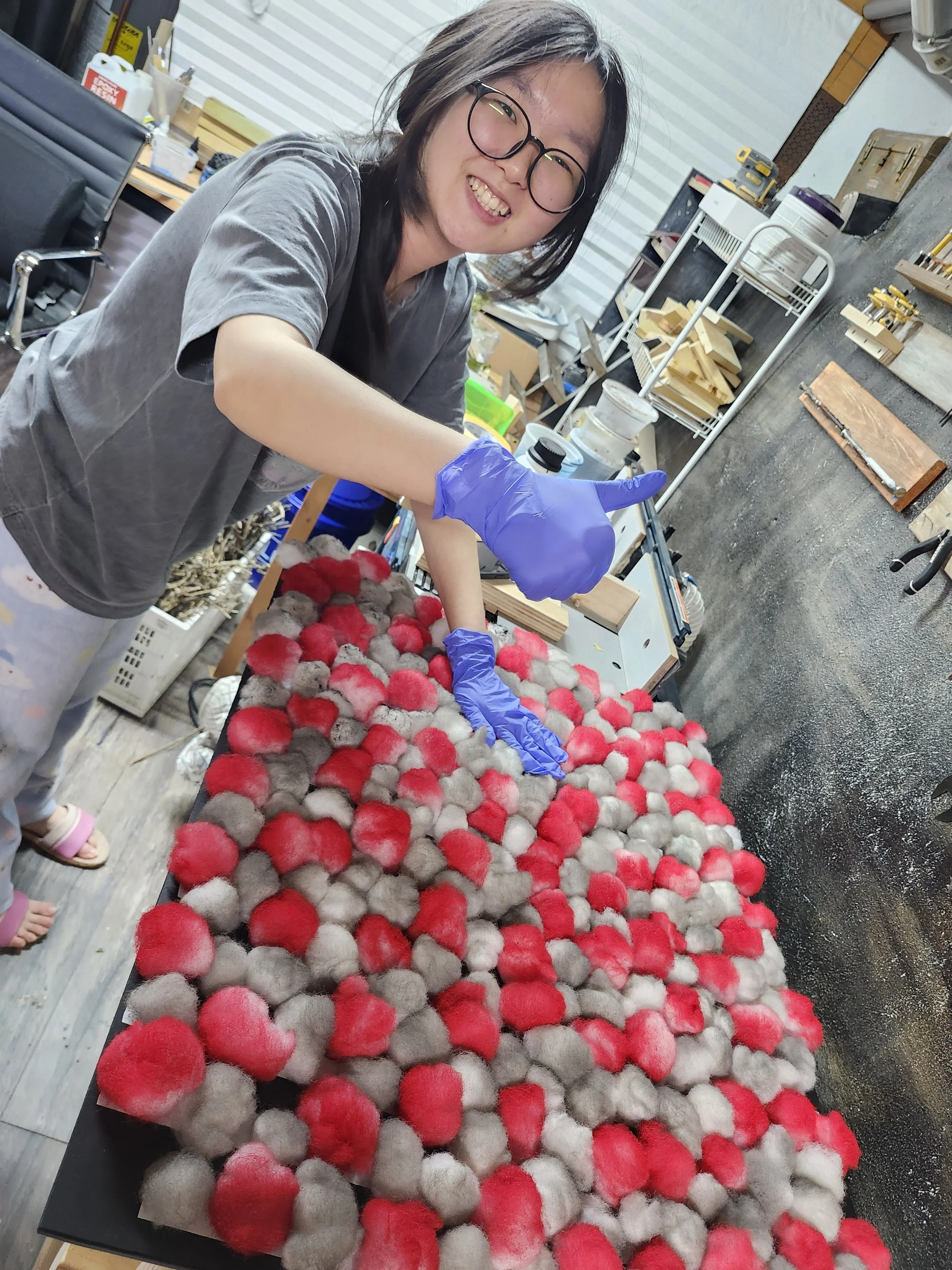 A woman wearing glasses and purple gloves is smiling and working with a large collection of colorful dyed fox fur puffs in a workshop. She is giving a thumbs-up gesture.