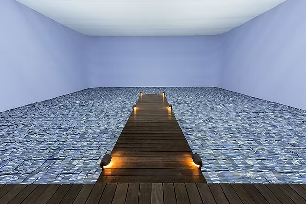 Indoor swimming pool with a wooden dock and lights at the end, enclosed by plain walls