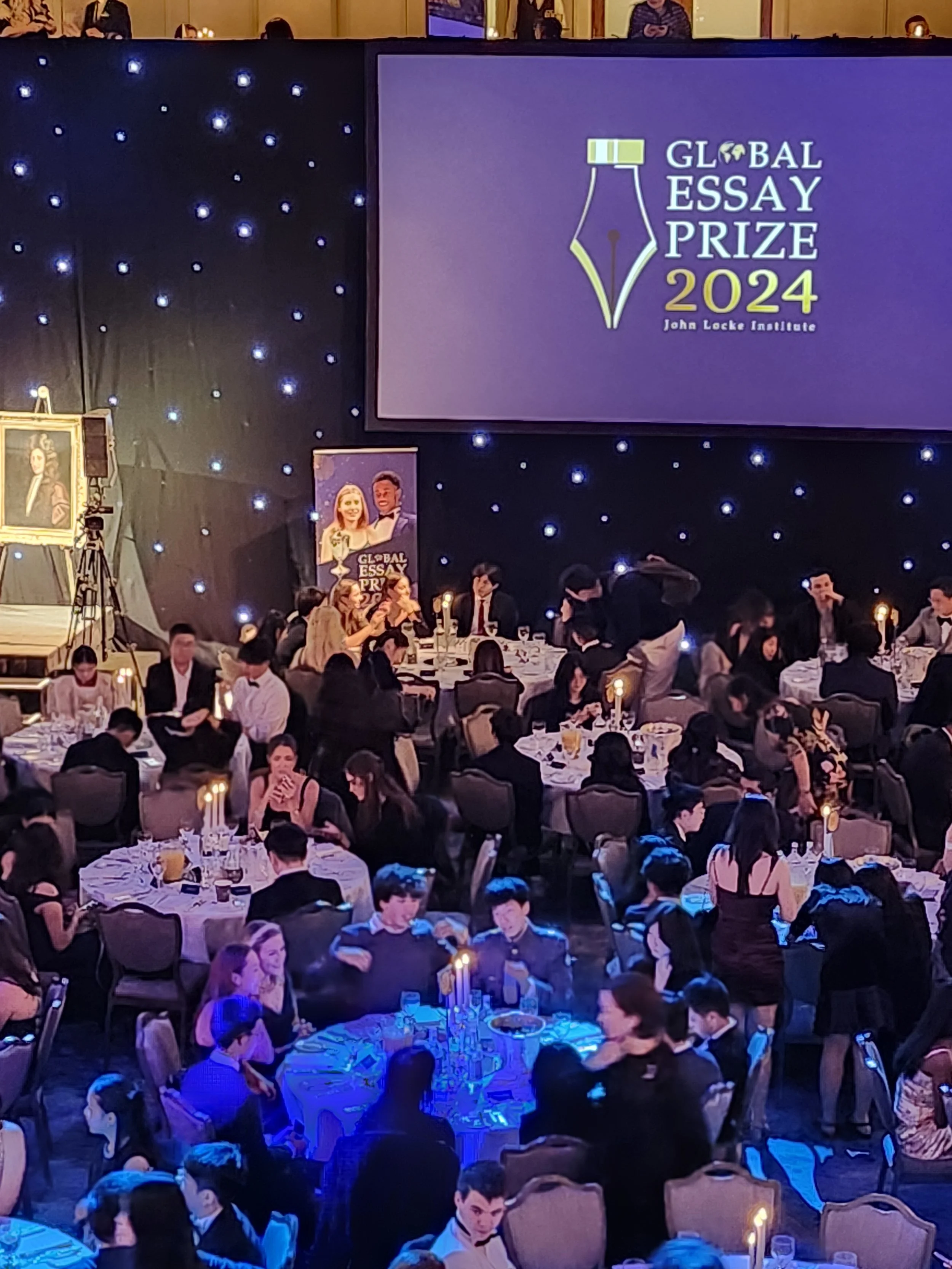 A large banquet hall filled with people seated at round tables, with some standing and talking. There is a large screen displaying 'Global Essay Prize 2024 John Locke Institute,' and a poster with two illustrated people, along with framed portraits on easels and star-like lights on the dark backdrop.