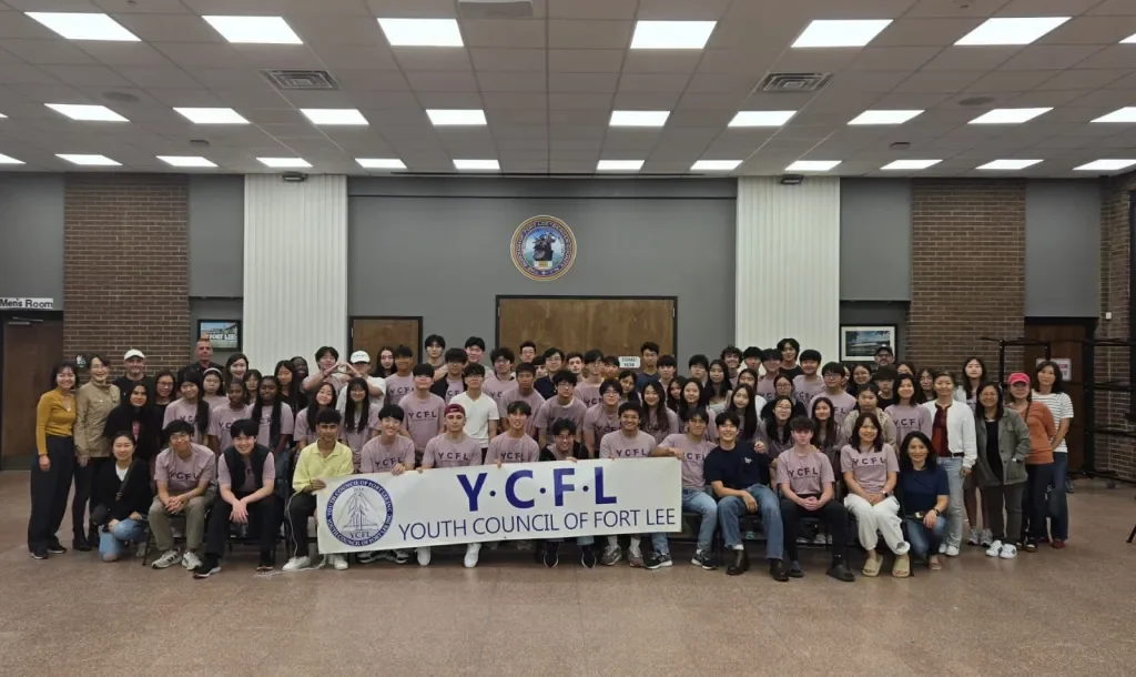 Group photo of youth members of the Youth Council of Fort Lee, gathered in a large indoor room, holding a banner with the organization's name.