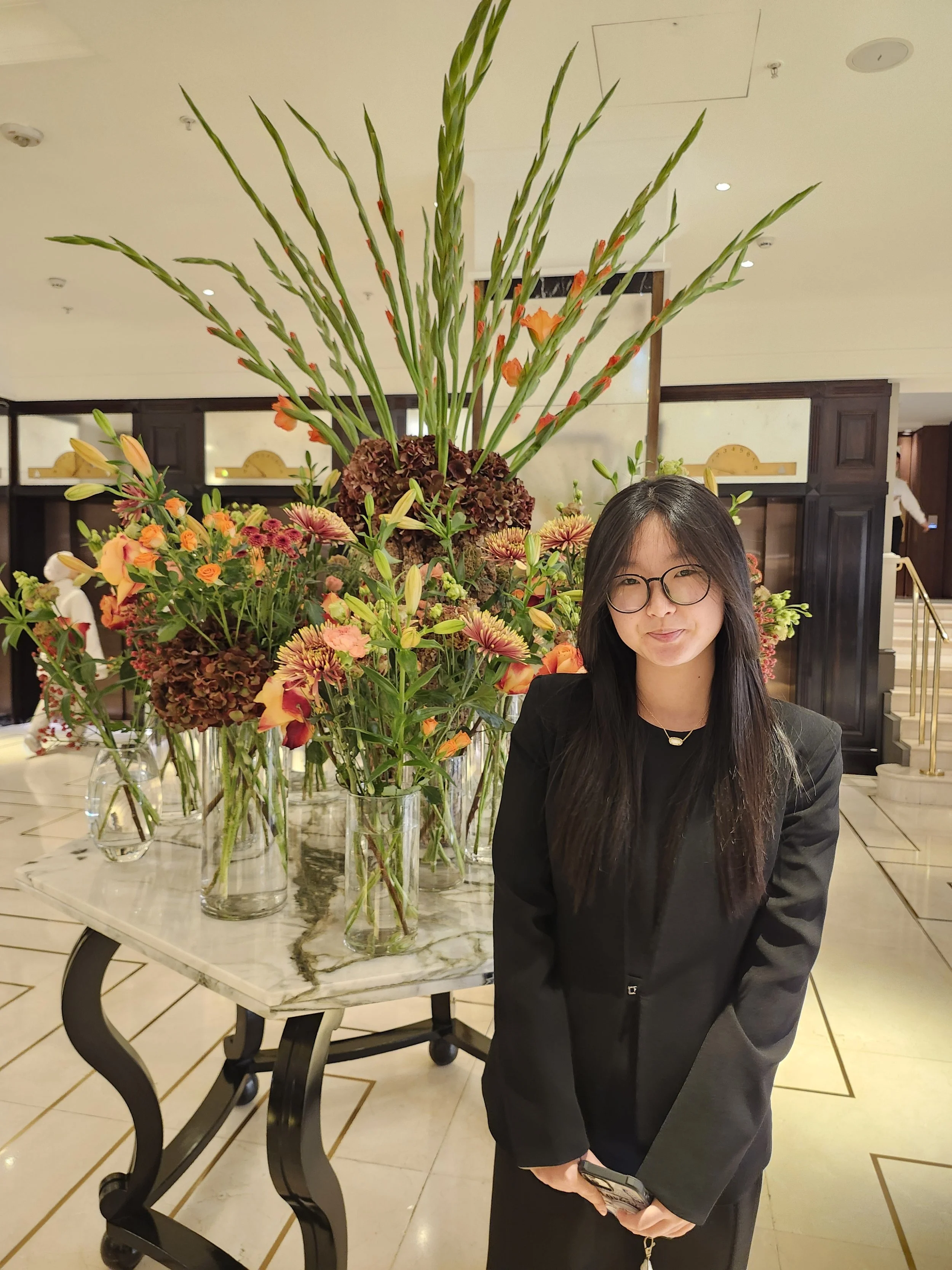 A young woman with glasses and long dark hair standing in front of a large floral arrangement in a hotel lobby.