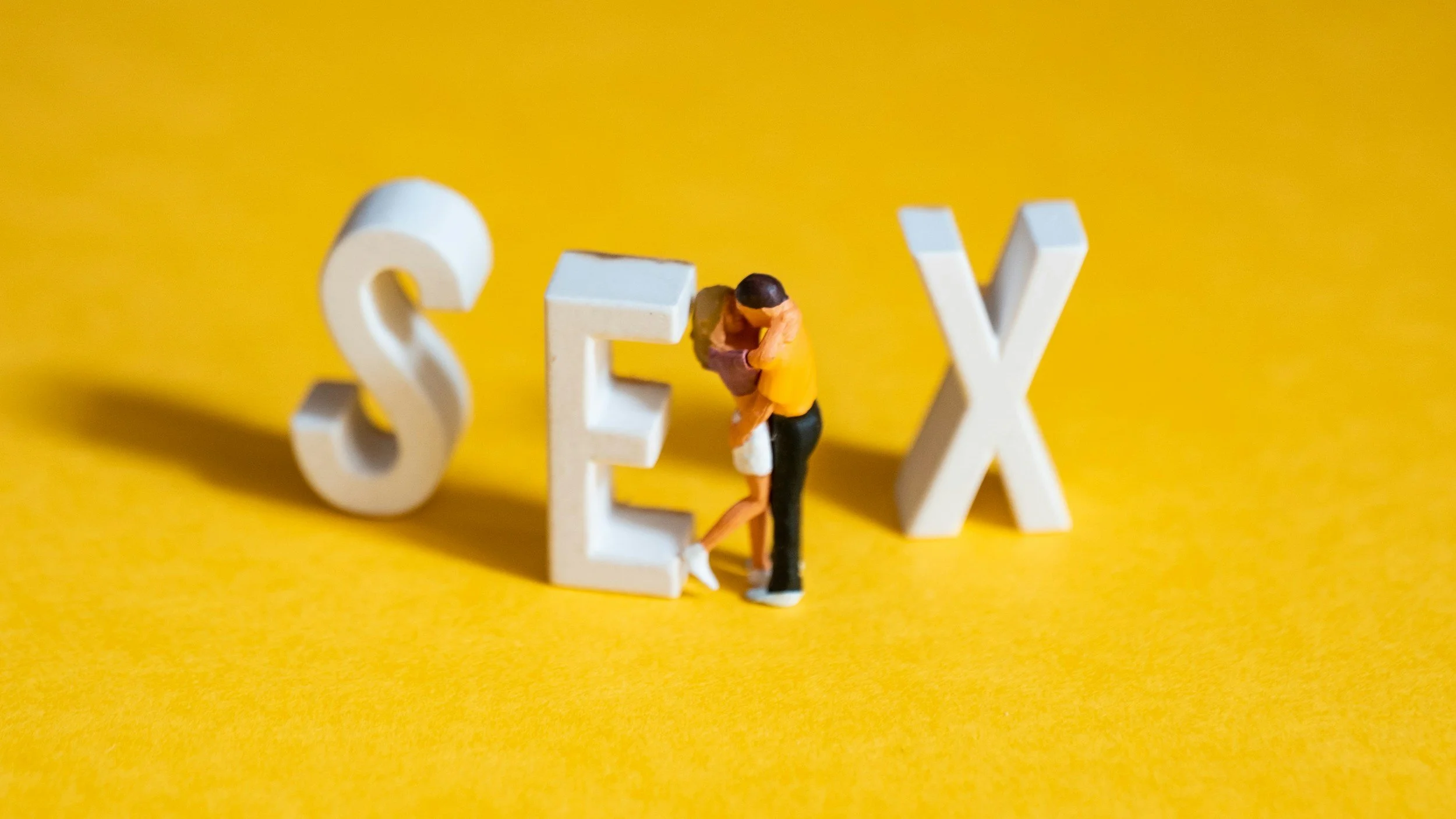 Miniature couple kissing next to large white letters spelling 'SEX' on a yellow background.
