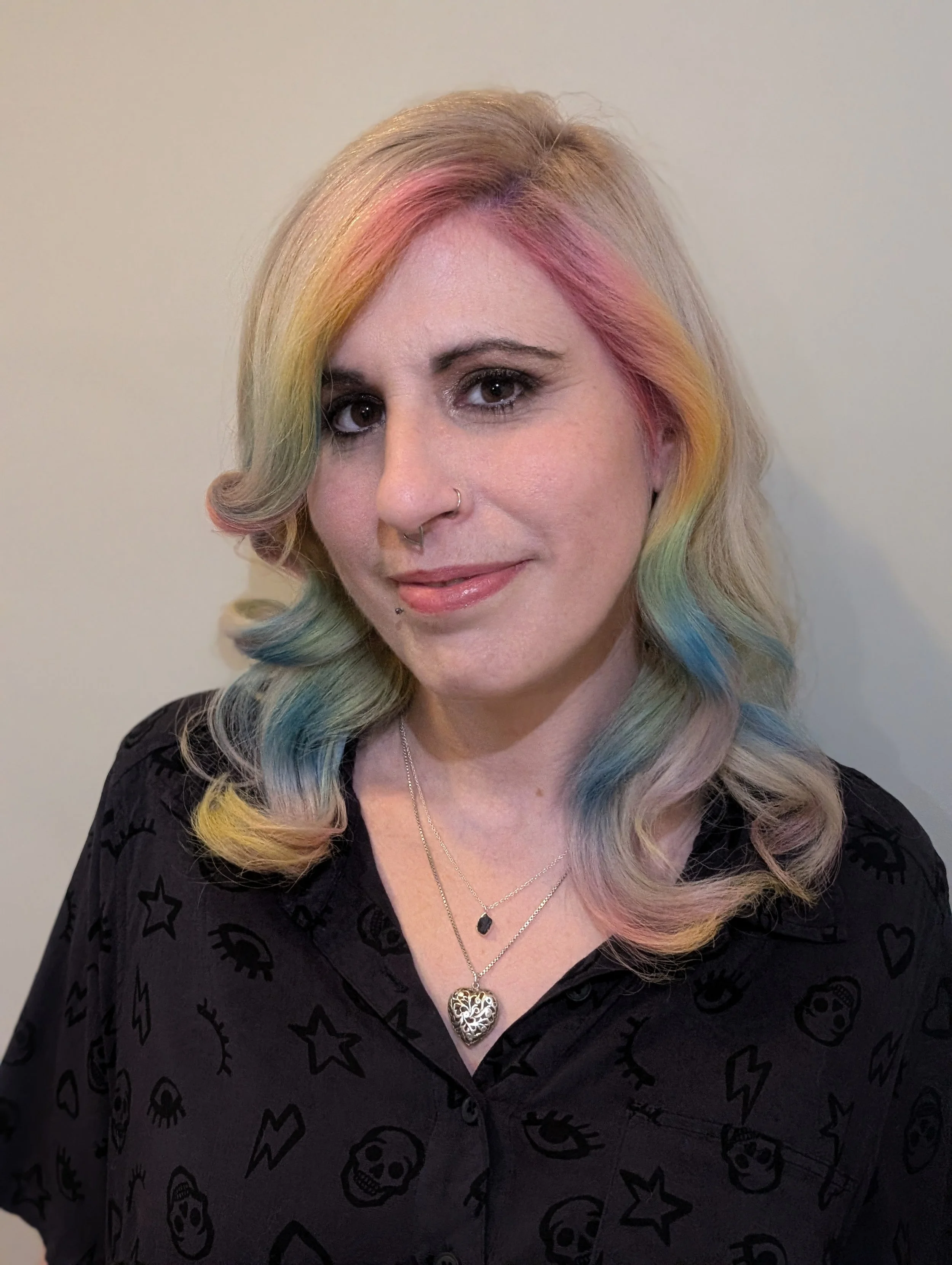 A woman with light skin and colorful, wavy hair featuring pastel shades of pink, yellow, green, and blue. She has a nose ring, a lip piercing, and is wearing layered necklaces and a black shirt with a pattern of skulls, stars, and eyes.