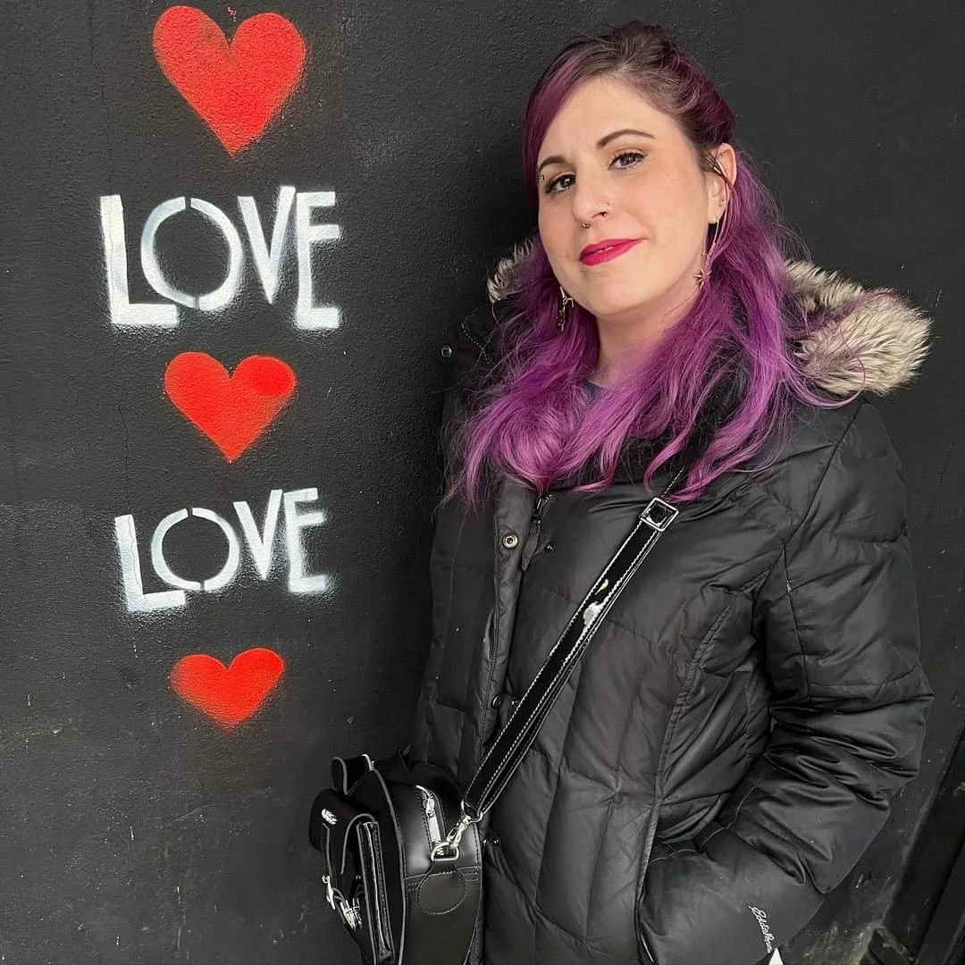 A woman with purple hair standing next to a black wall with the word 'LOVE' written twice and three red hearts painted on it.