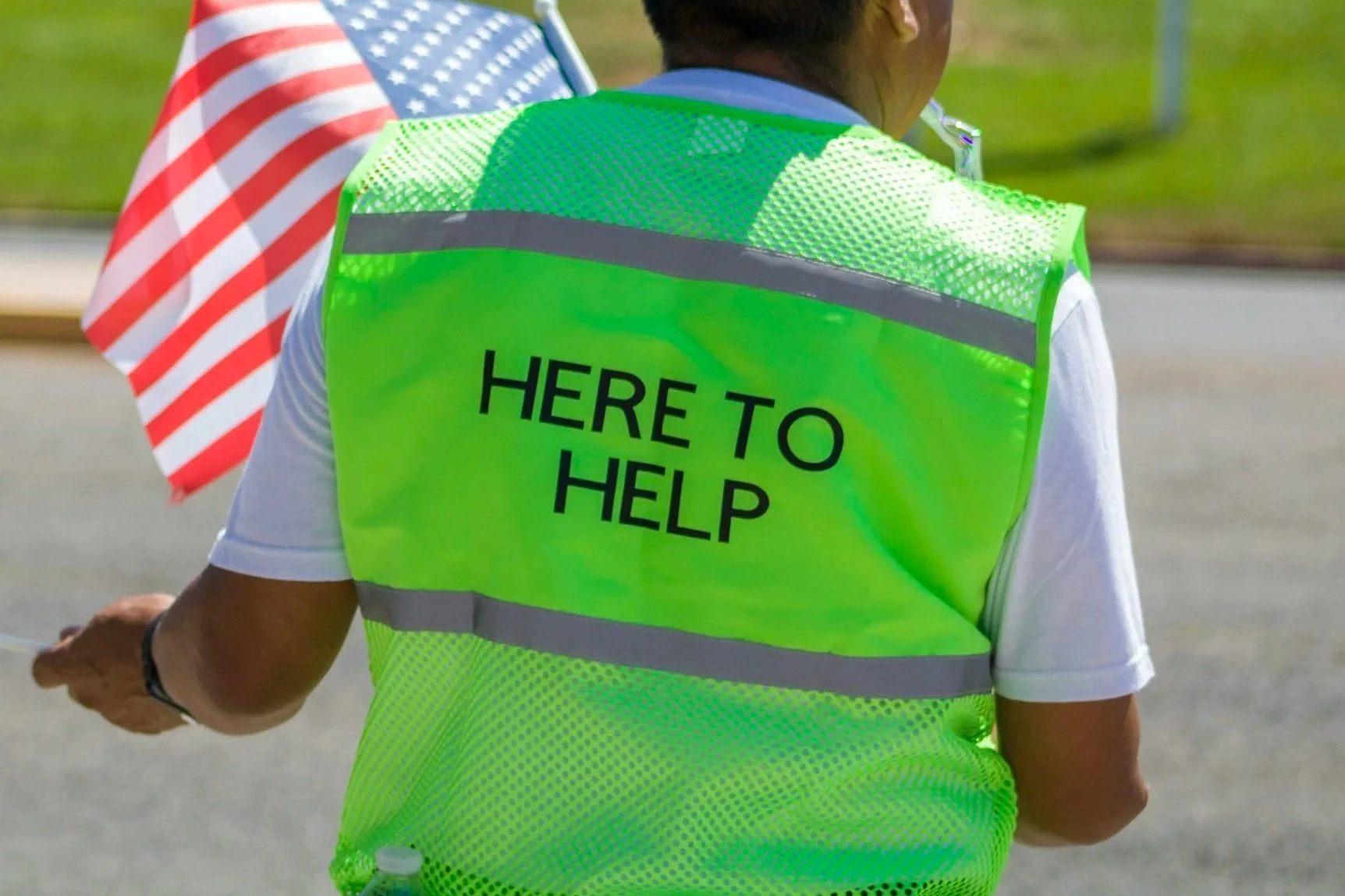 A person wearing a neon green safety vest with the words "HERE TO HELP" printed on the back, standing outdoors near a road.