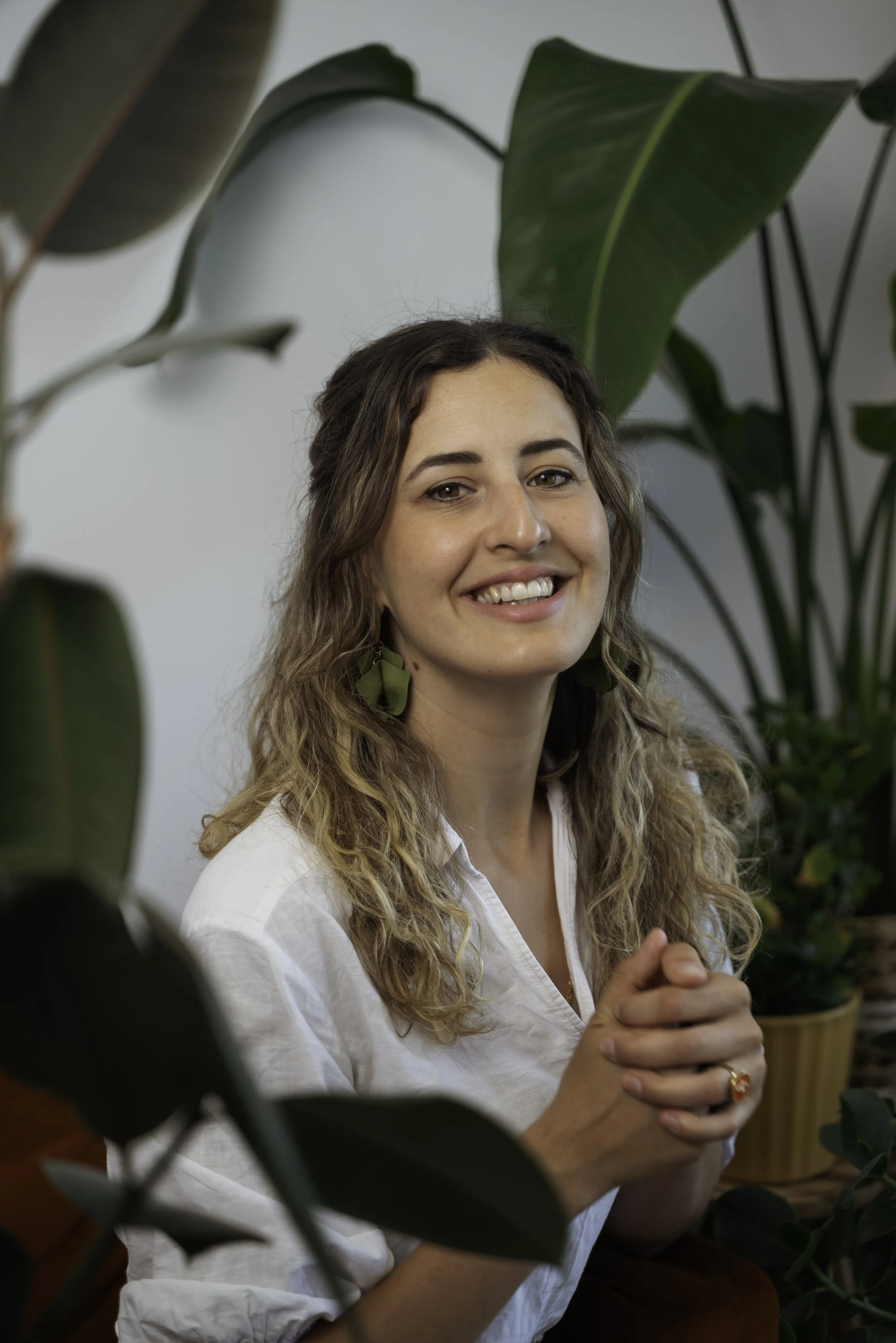 A woman with long wavy hair, wearing a white shirt and green earrings, smiling and sitting among large green indoor plants.