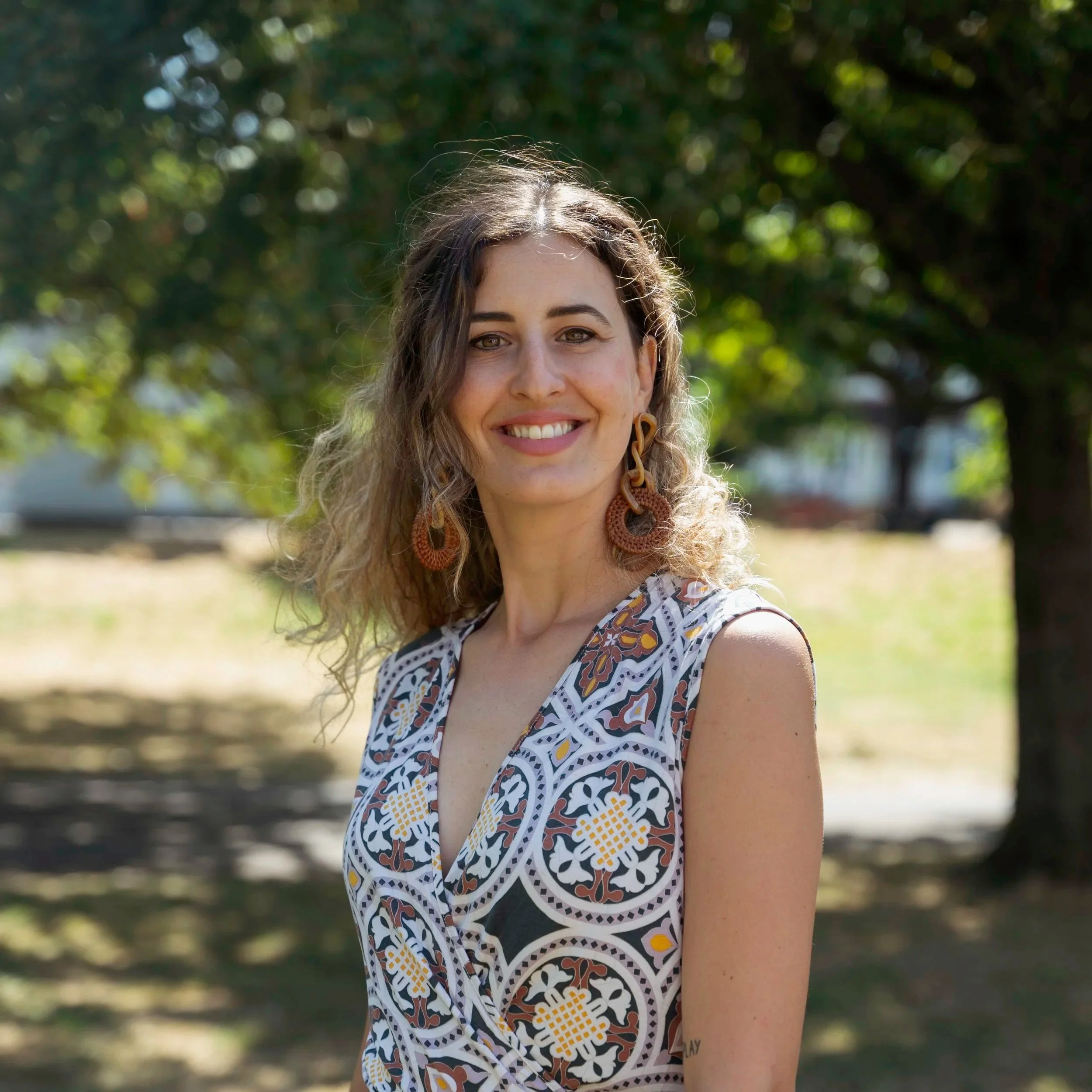 A woman with curly brown hair smiling outdoors on a sunny day, wearing a patterned sleeveless top and large earrings, with trees and greenery in the background.
