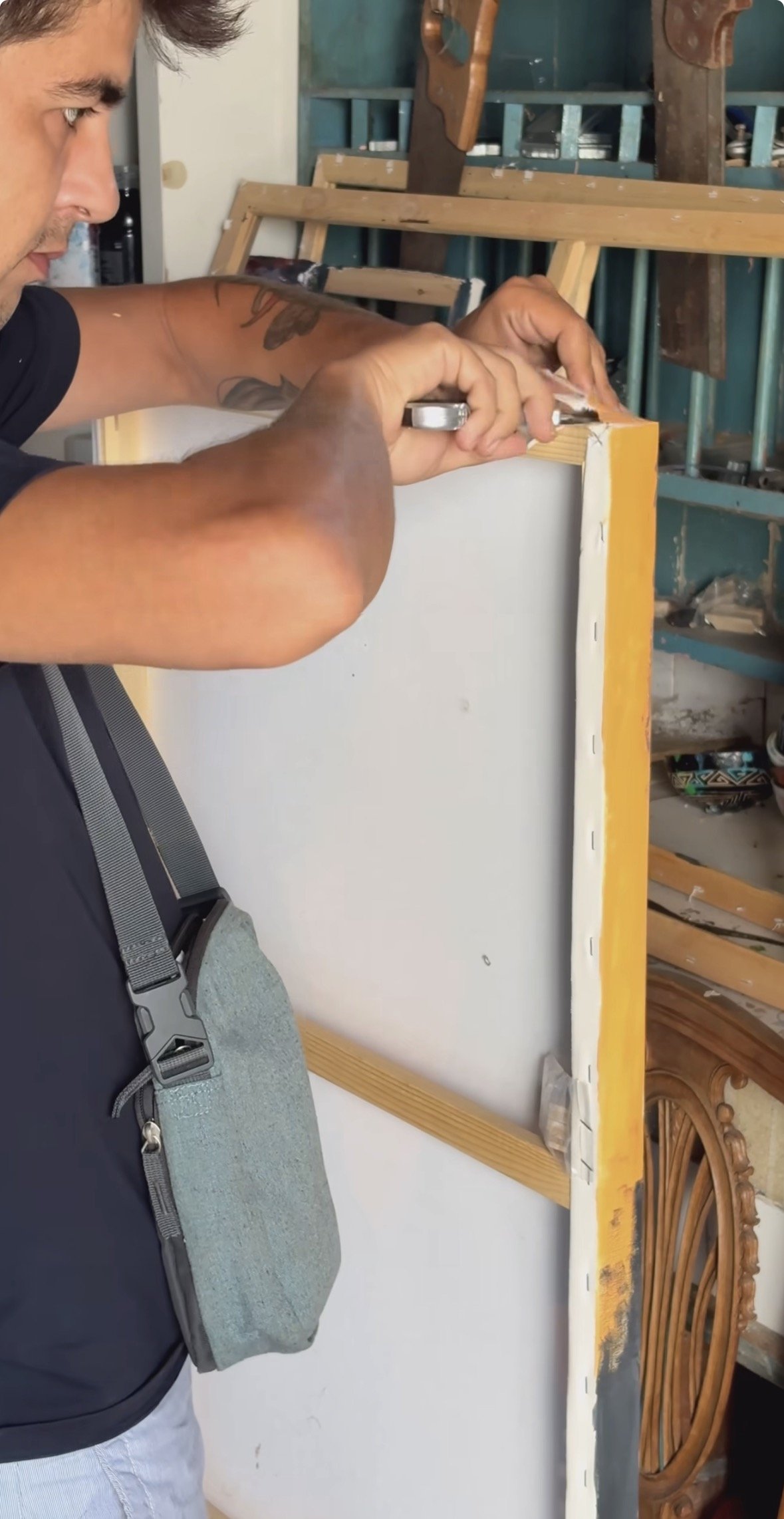 A man with tattoos is drilling into a wooden structure in a workshop. The workshop has tools and a wooden wheel visible in the background.