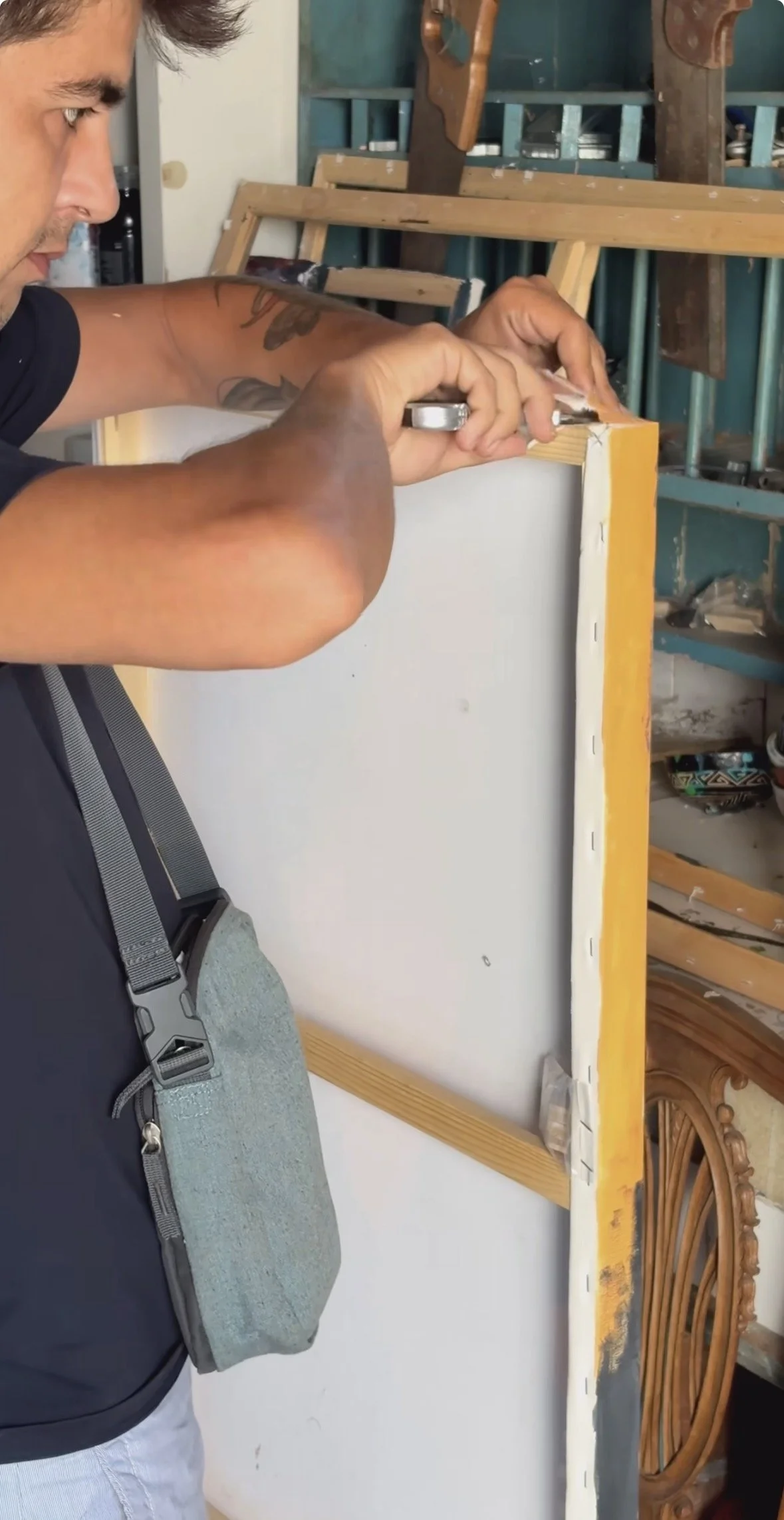A man with tattoos is drilling into a wooden structure in a workshop. The workshop has tools and a wooden wheel visible in the background.
