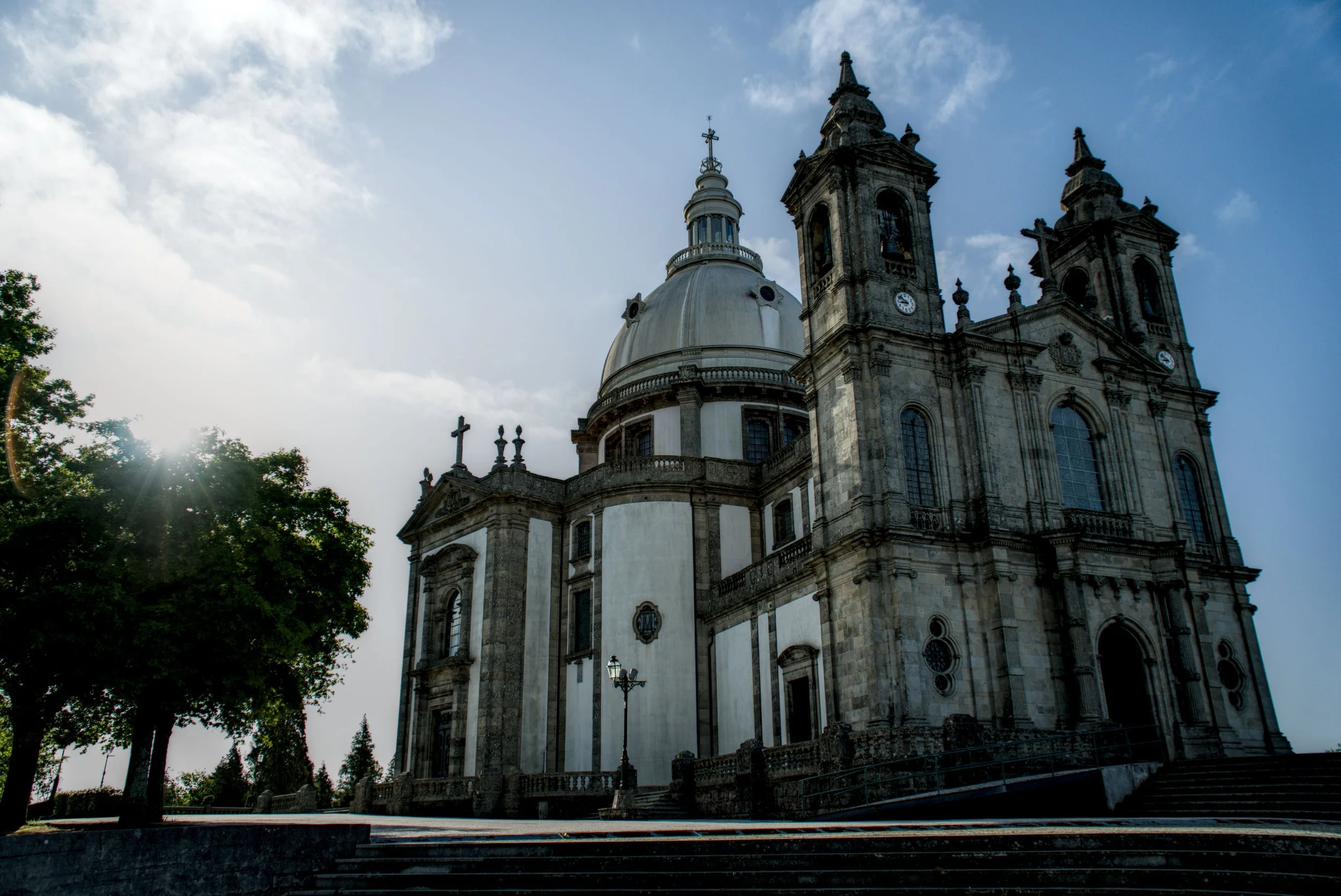 A historic church with twin bell towers and a large dome, set against a blue sky with some clouds, surrounded by trees and street lamps.