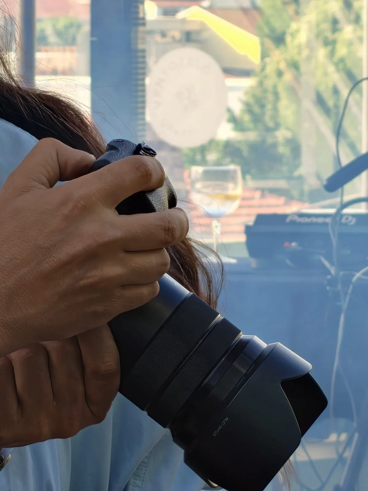 A person holding a camera with a large telephoto lens, pointing it through a glass window with a blurred outdoor scene in the background.