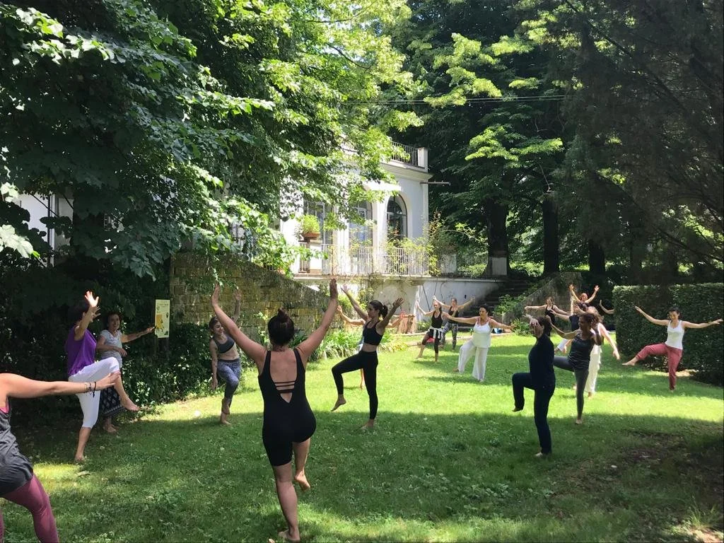 Gruppo di persone che praticano yoga all'aperto in un giardino con molti alberi e un edificio bianco sullo sfondo.