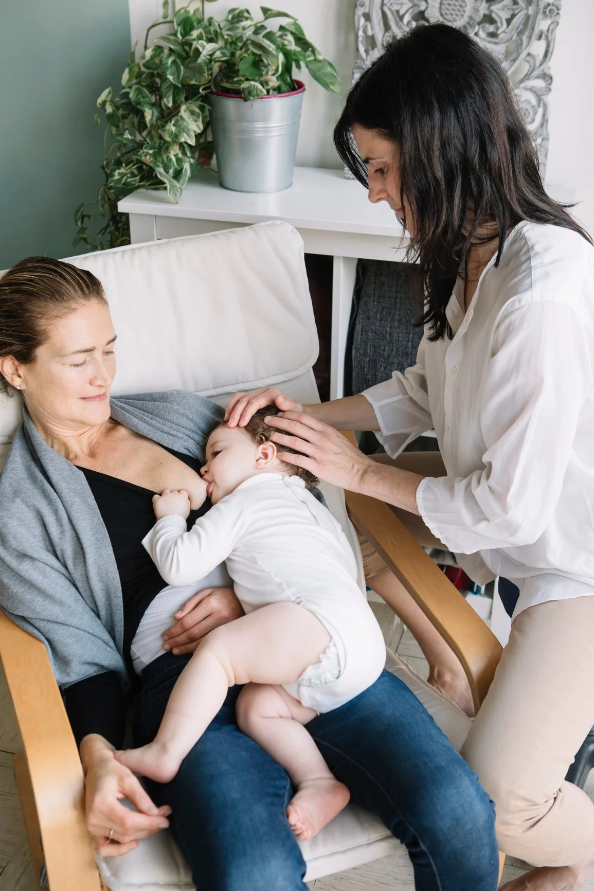 A woman breastfeeding a young child while sitting on a chair, with another woman gently touching the child's head, in a cozy room with a plant on a white table in the background.