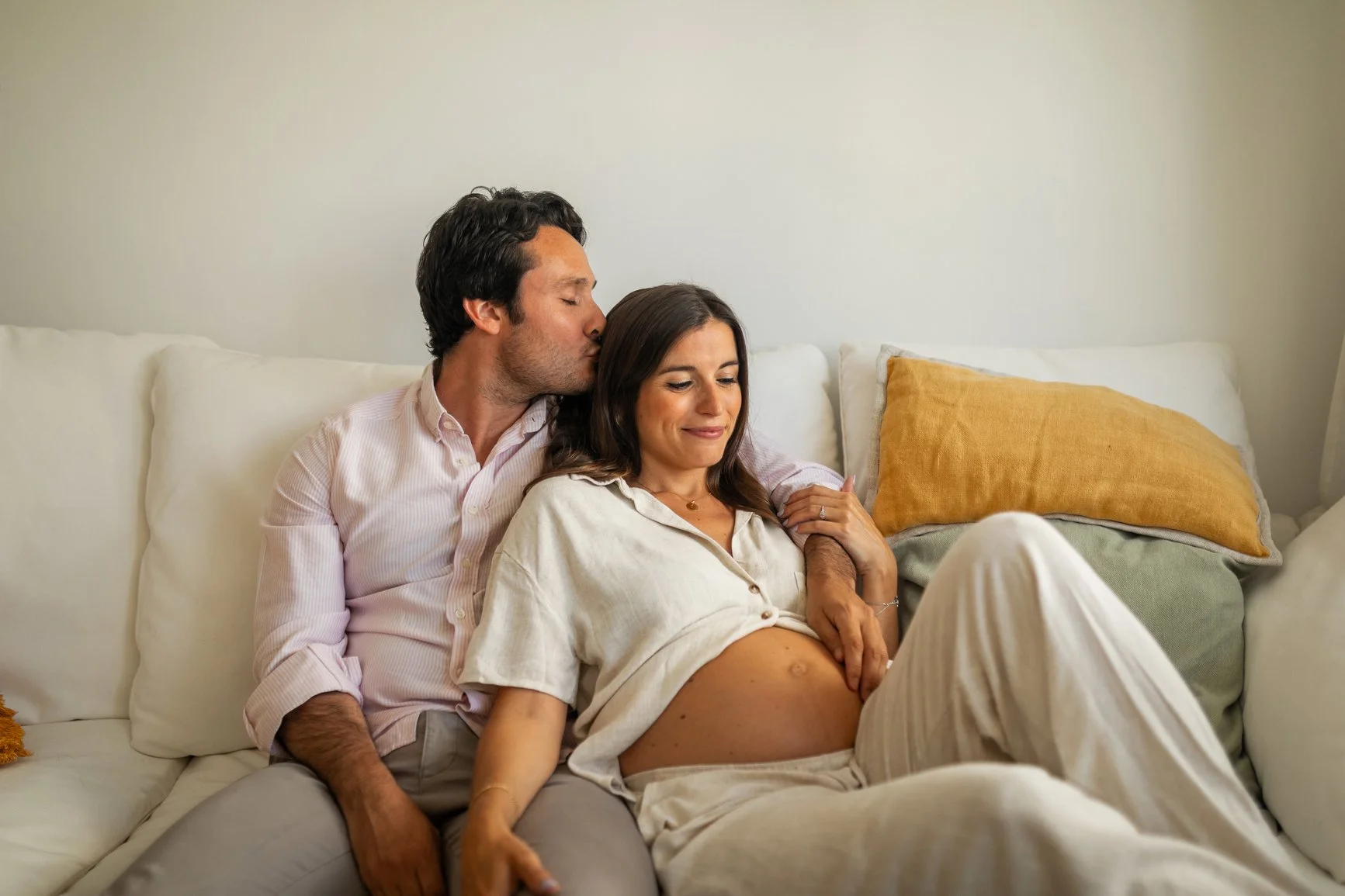 A man kissing a pregnant woman on the forehead while sitting on a white sofa in a cozy living room.
