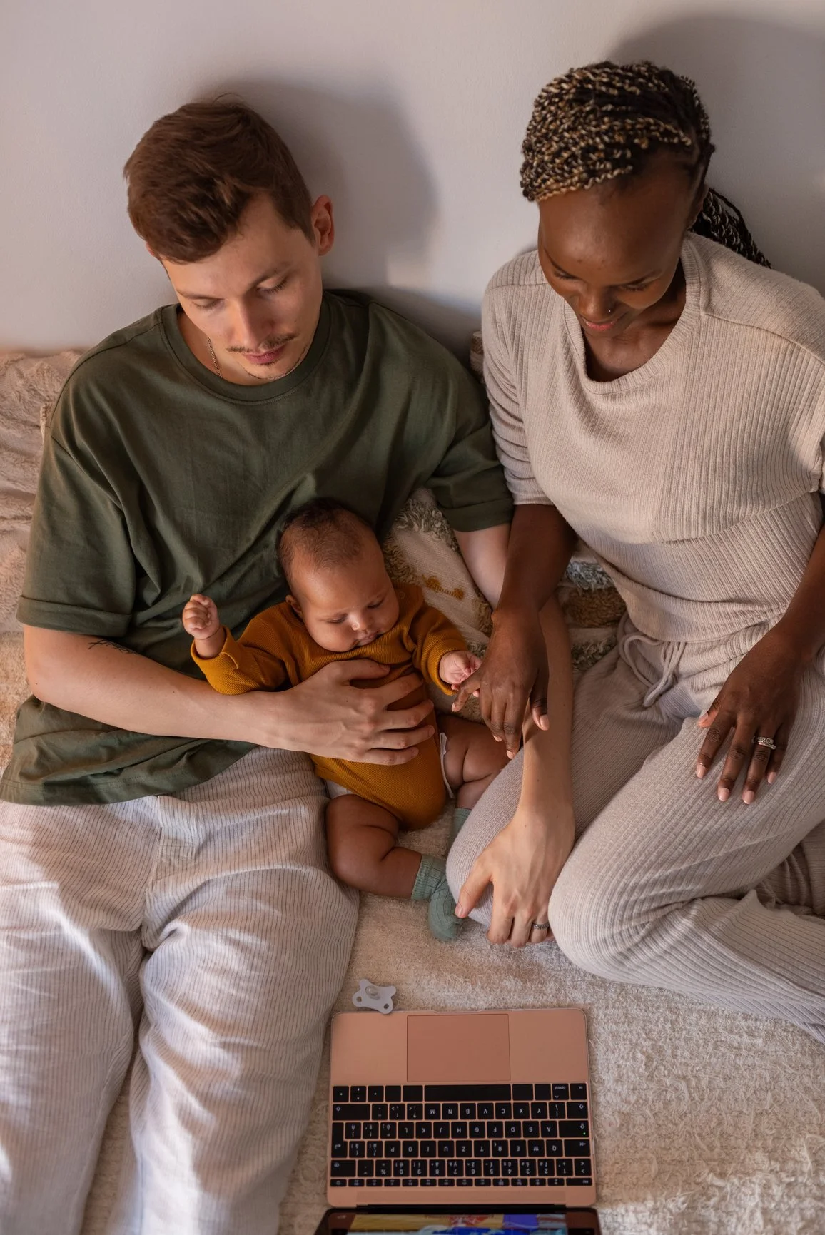 A family sitting on a bed, with an infant in the father's lap, using a laptop on the bed, sharing a tender moment.