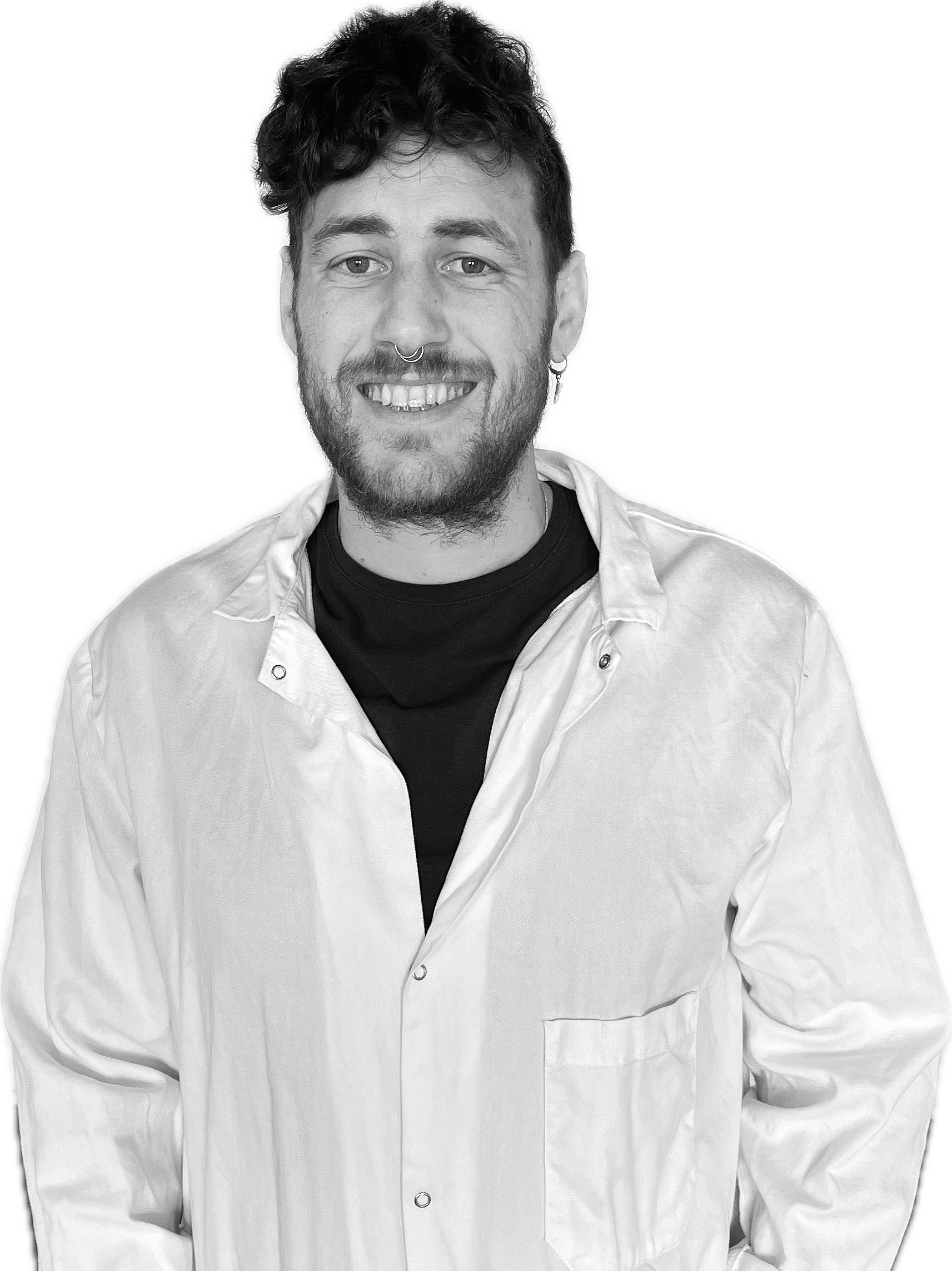 Black and white photo of a smiling man with curly hair, a beard, wearing a lab coat over a black shirt, standing against a black background.