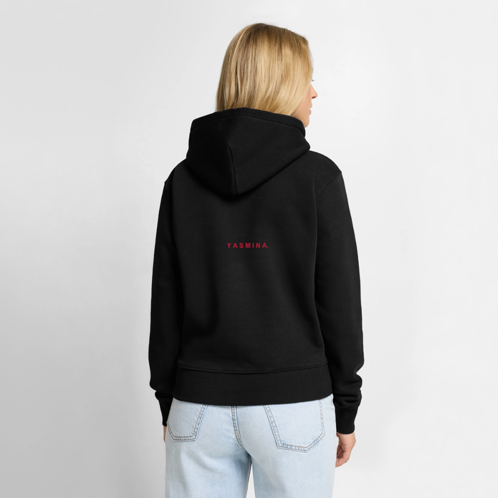En kvinde med blond hår iført en sort hoodie med teksten 'YASMINA.' på ryggen, set bagfra, mod en hvid baggrund.
