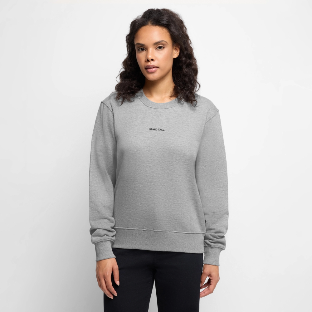 Kvinde med mørkt, krøllet hår iført grå sweatshirt med teksten 'STAND TALL.' og sorte bukser, står foran en hvid baggrund.