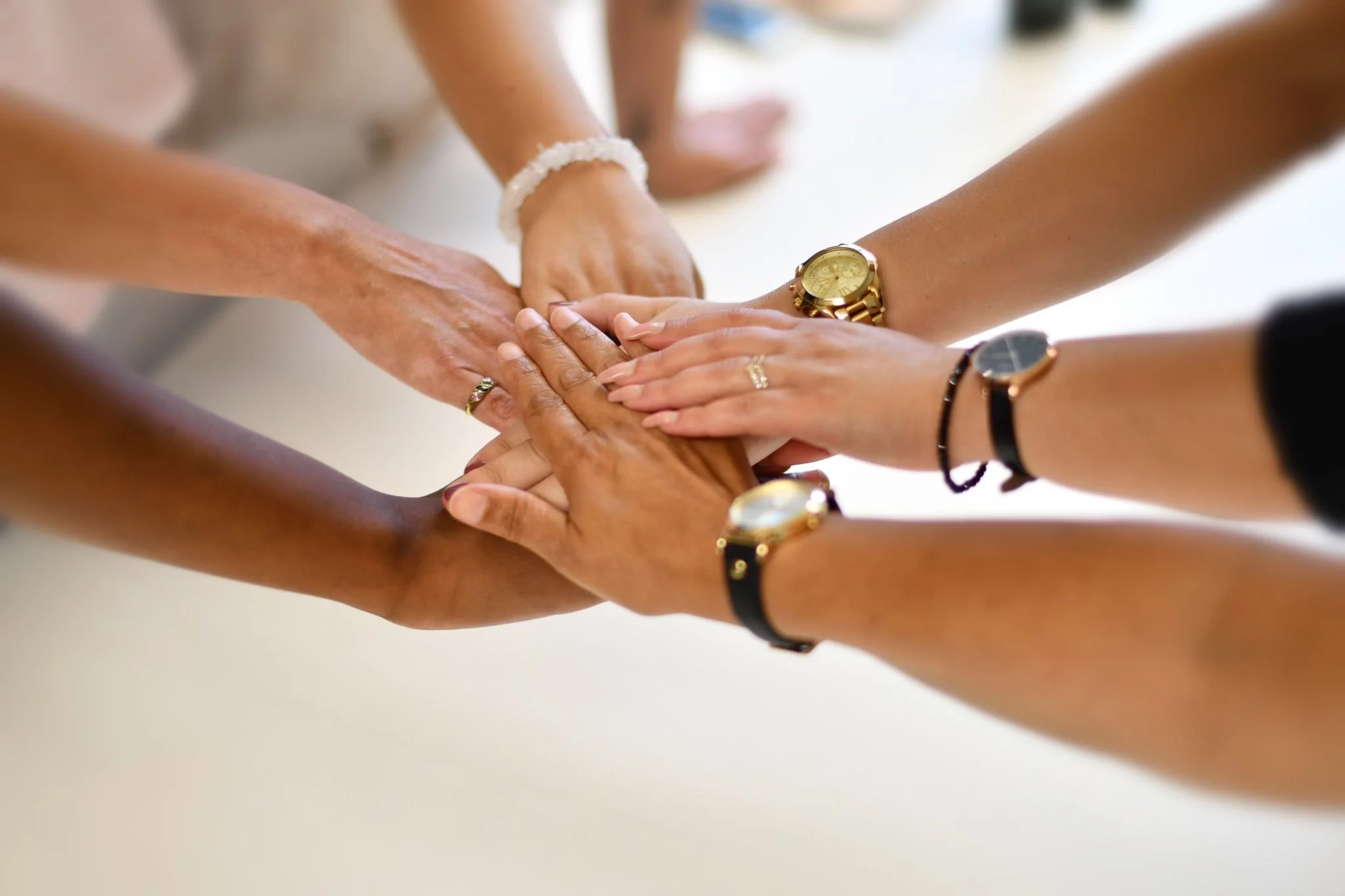 Multiple hands with different skin tones stacked on top of each other, wearing various watches and rings, symbolizing teamwork or unity.