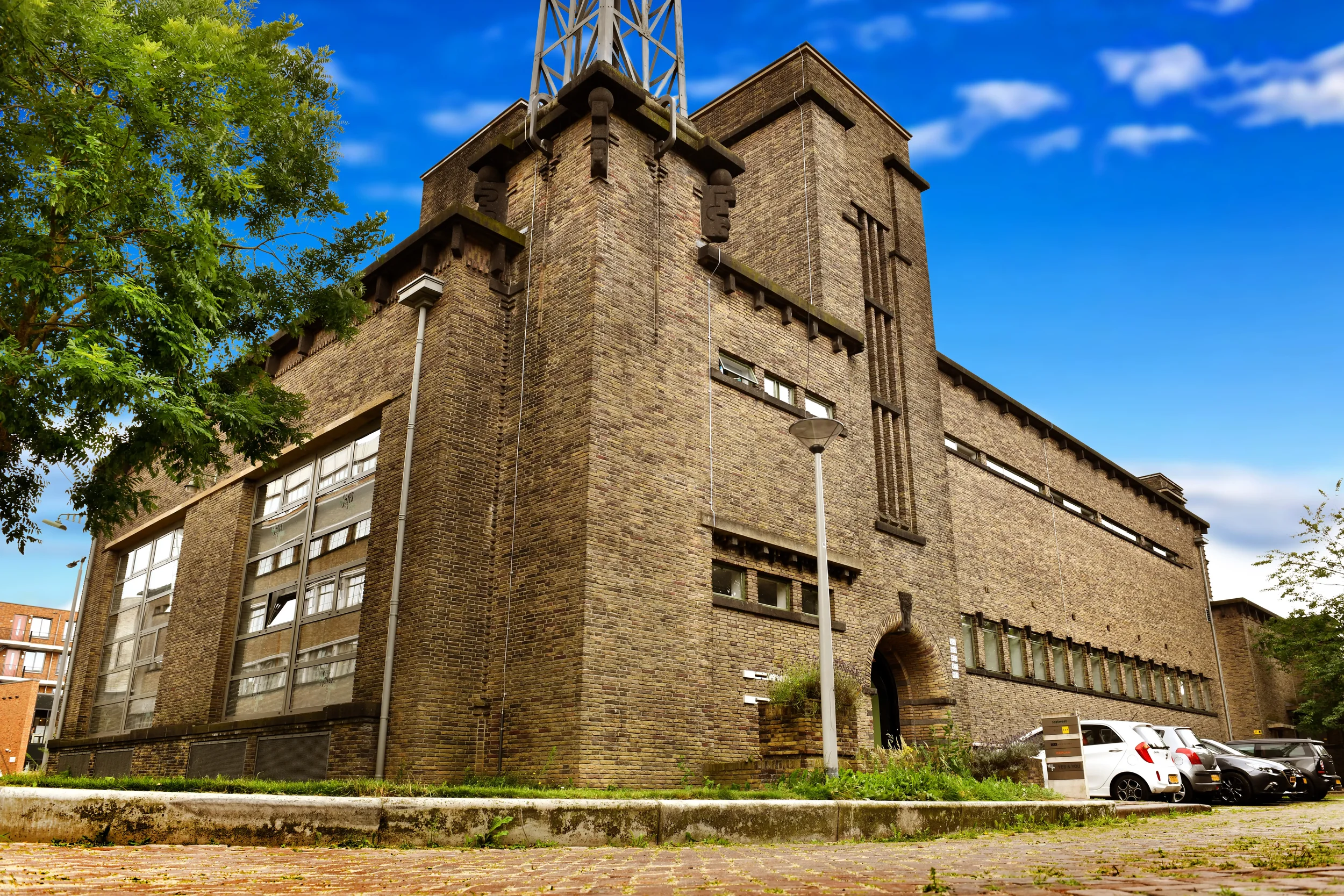 View of a multi-story brick building with rectangular windows, a small archway entrance, and a tall metallic tower structure on top. There are trees with green leaves to the left, a parking lot with several cars at the bottom, and a clear blue sky with some clouds in the background.