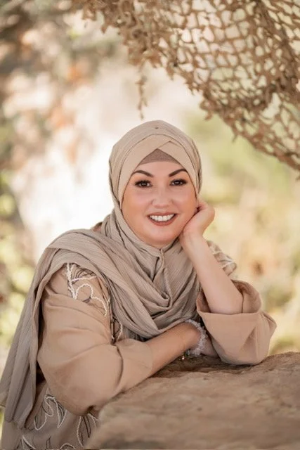A woman with a head wrap smiling at a desk with a computer and a cup, standing in an office with a window and a wall with decorative swords.