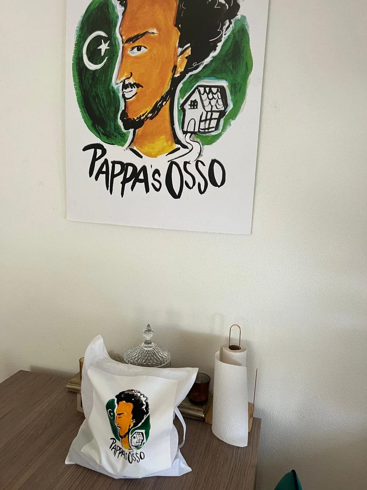 A poster and a tote bag with a drawing of a man with dark hair and a goatee, script that says "Pappas Osso," and a small house illustration. The poster and bag are on a wooden table against a plain white wall.