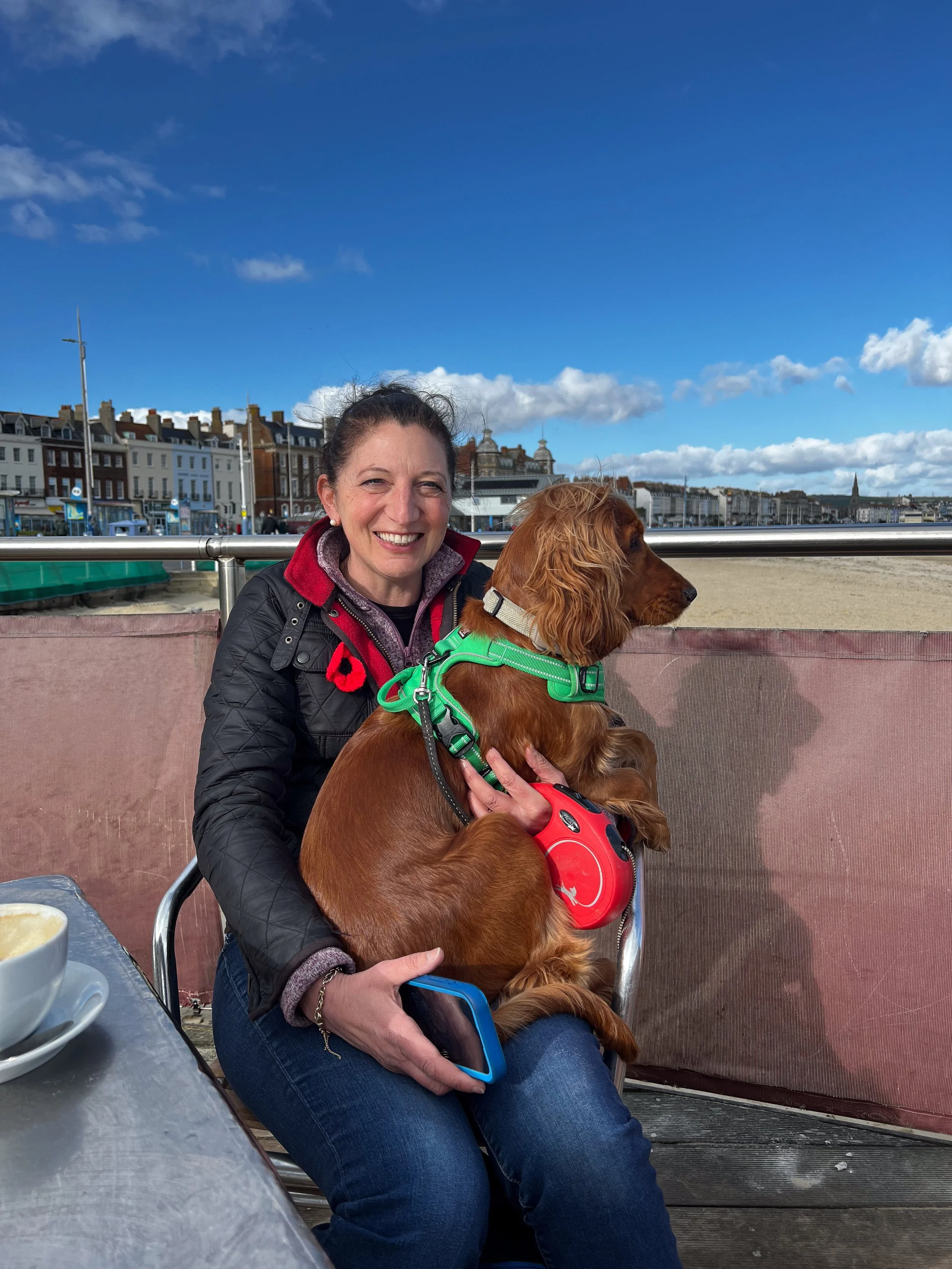 A woman smiling while holding a brown spaniel dog in a green harness, seated outdoors at a café table with a drink, with a beach, colorful buildings, and a blue sky with clouds in the background.