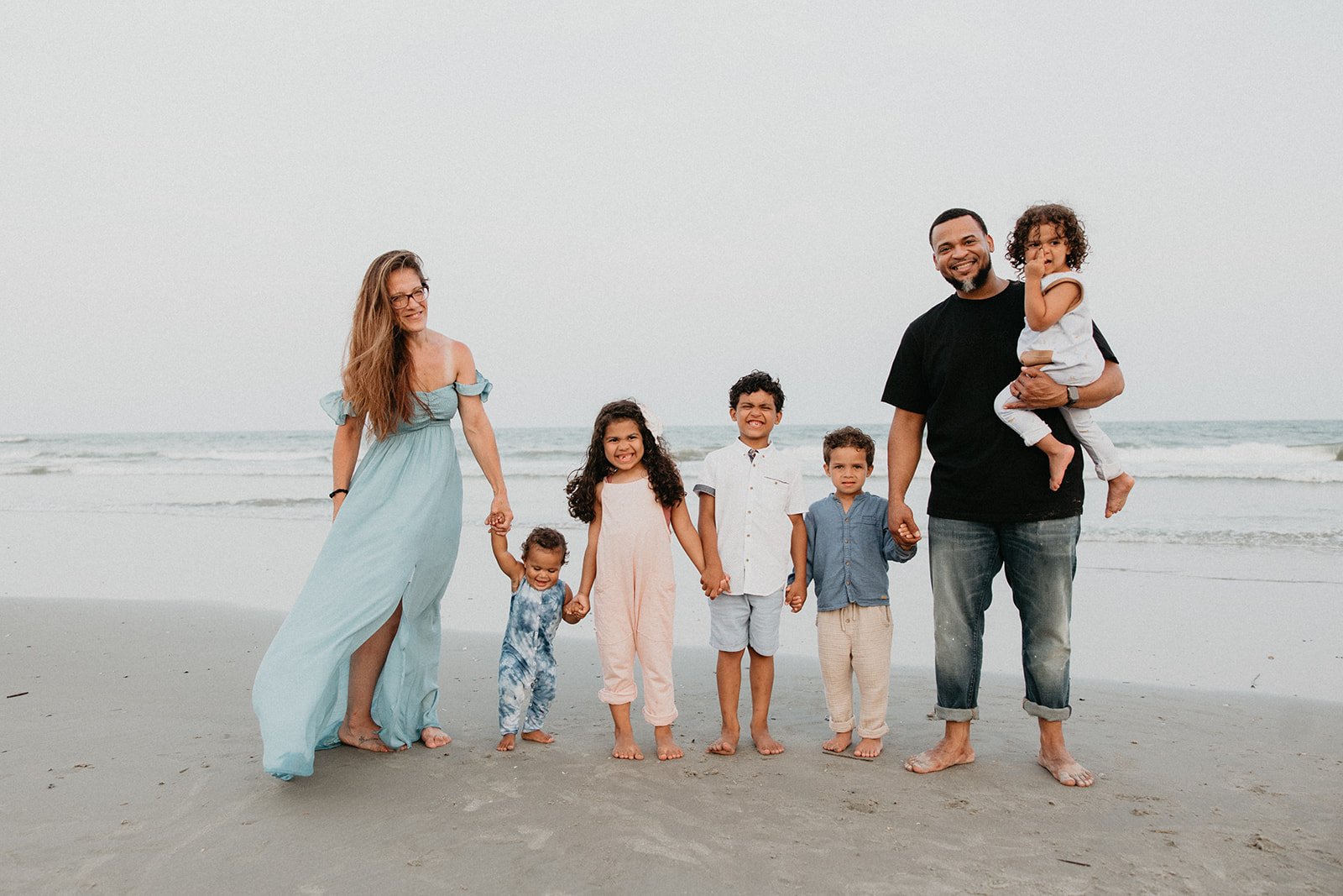 A diverse family of eight, including adults and children, walking along the beach holding hands and smiling, with the ocean in the background.