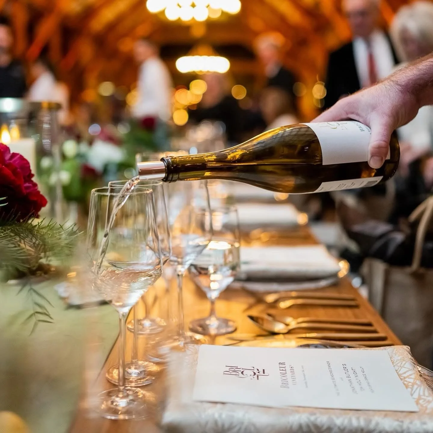 A person pouring white wine into a glass at a formal dinner event, with multiple glasses and a decorated table in the background and blurred guests.