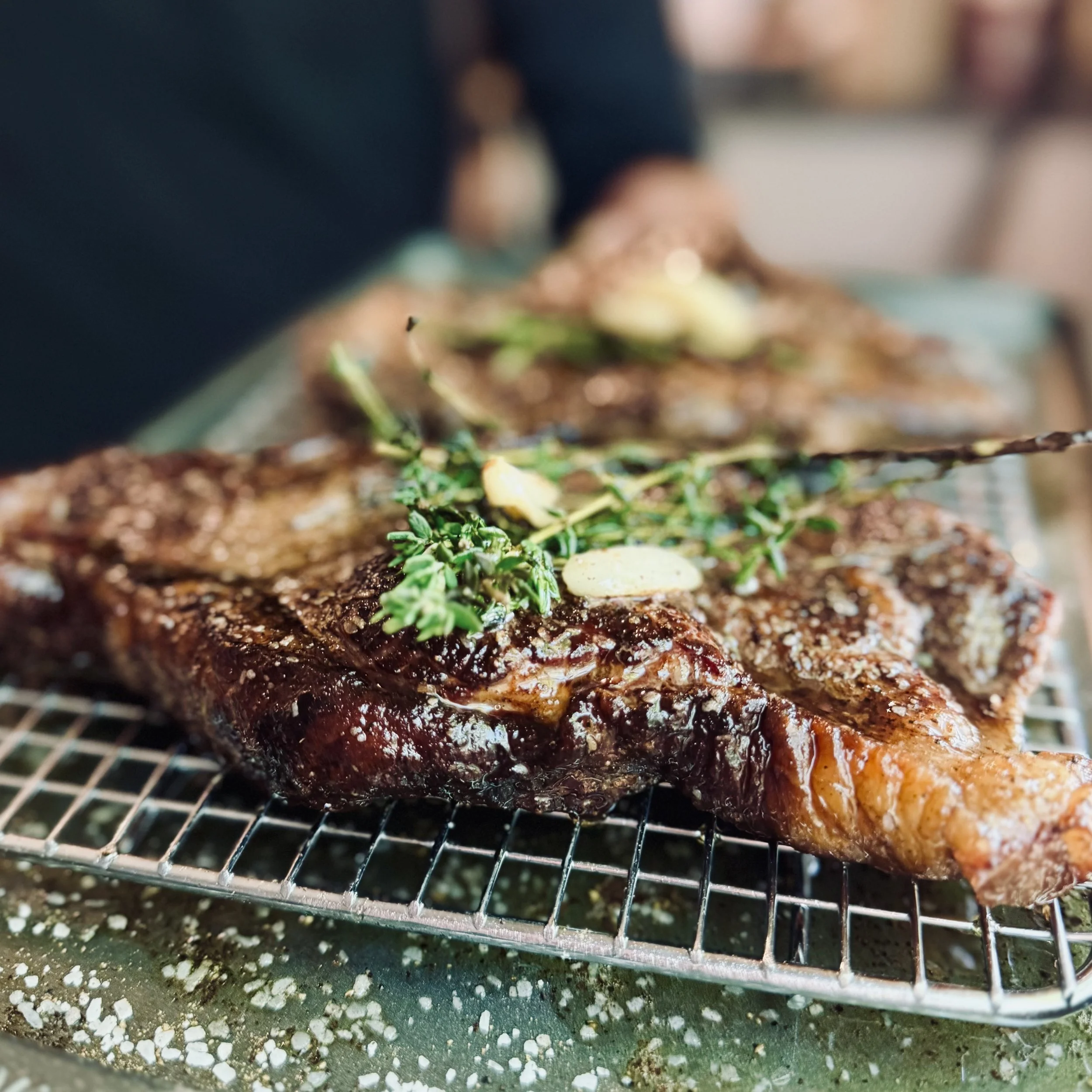 Close-up of a cooked meat dish garnished with herbs, cooling on a wire rack with scattered seasoning and crumbs beneath.