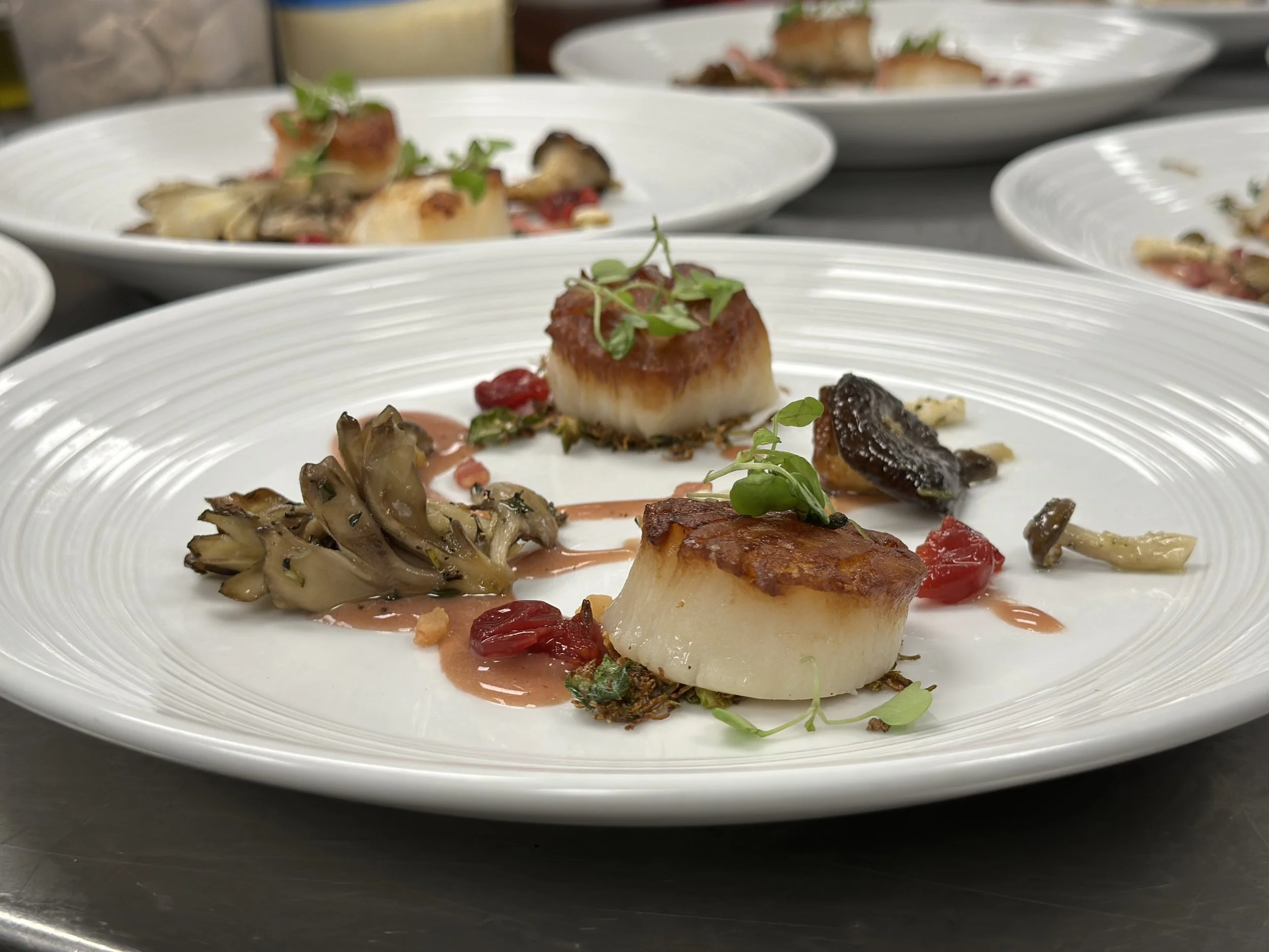 A white plate with a piece of cooked white fish garnished with microgreens, served with roasted mushrooms, roasted red peppers, and a drizzle of light pink sauce.