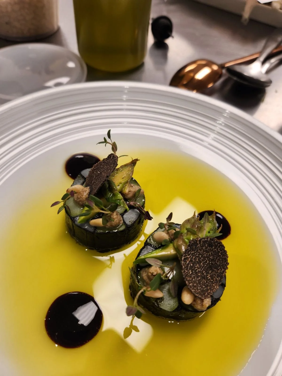 Two cylindrical servings of a gourmet dish with vegetables, garnished with microgreens, black truffle slices, and dark sauce, in a yellow oil-based liquid on a white plate.