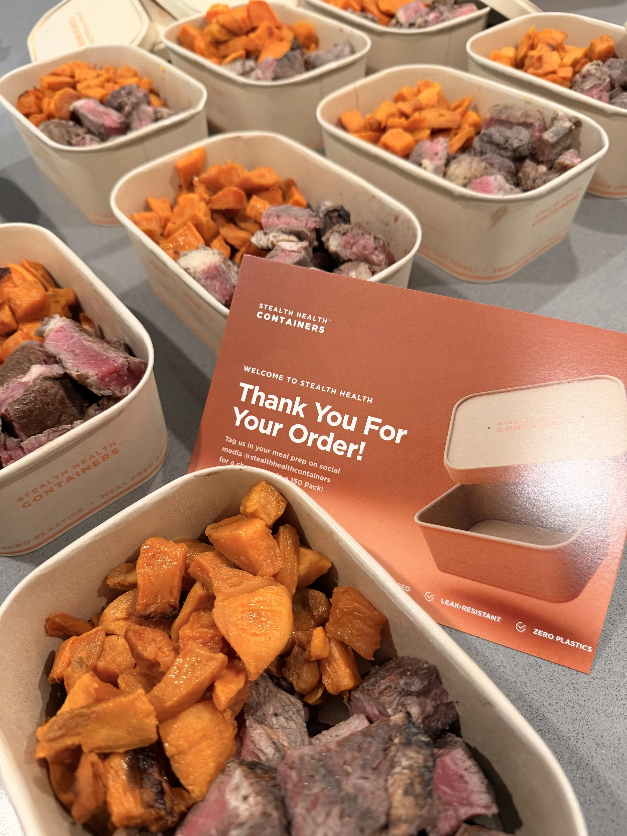 Multiple white containers filled with prepared cooked food, including roasted sweet potatoes and sliced steak, at a food service event.