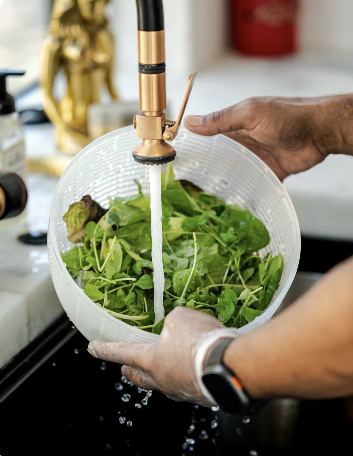 Person rinsing fresh green salad in a clear plastic bowl under a kitchen faucet.