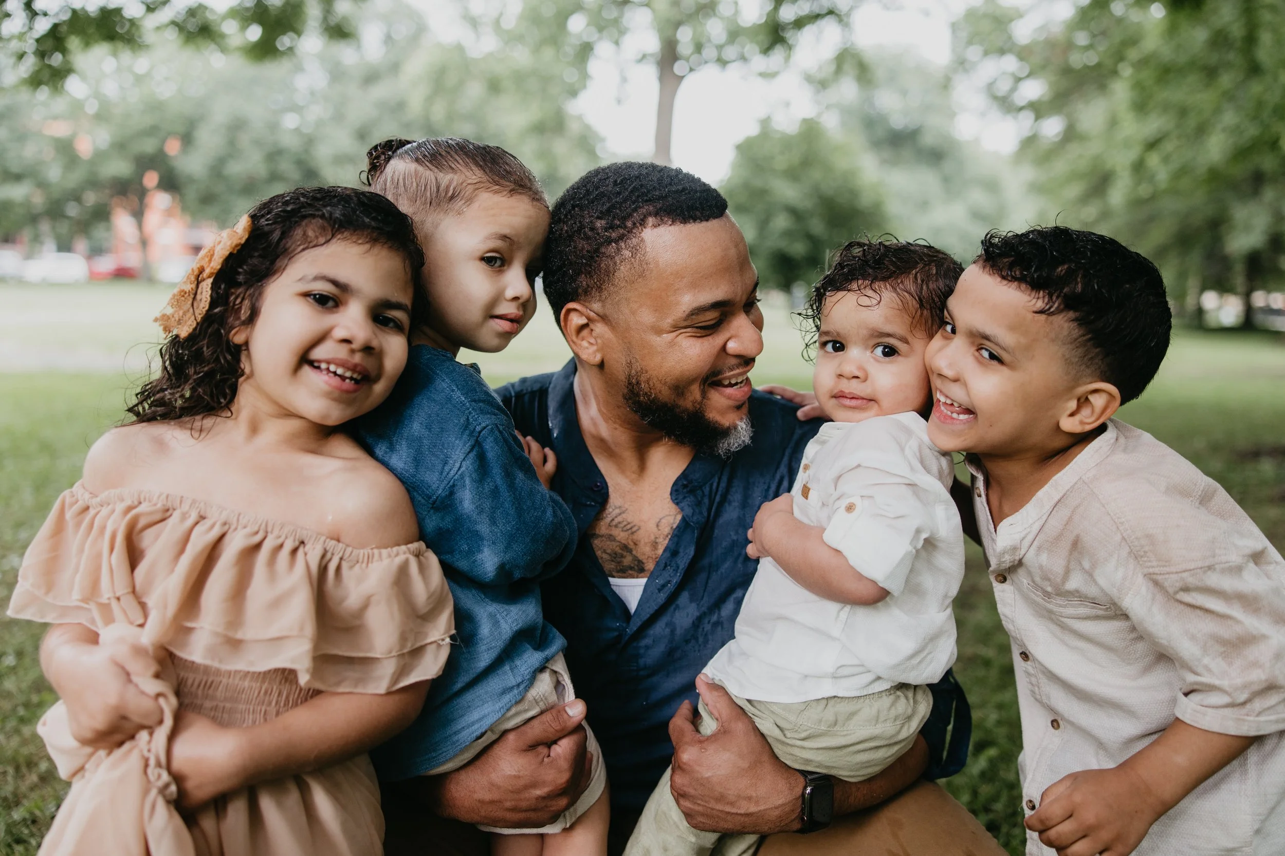 A man with tattoos smiling and holding four children of different ethnicities, all smiling and enjoying time together outdoors in a park surrounded by green trees.