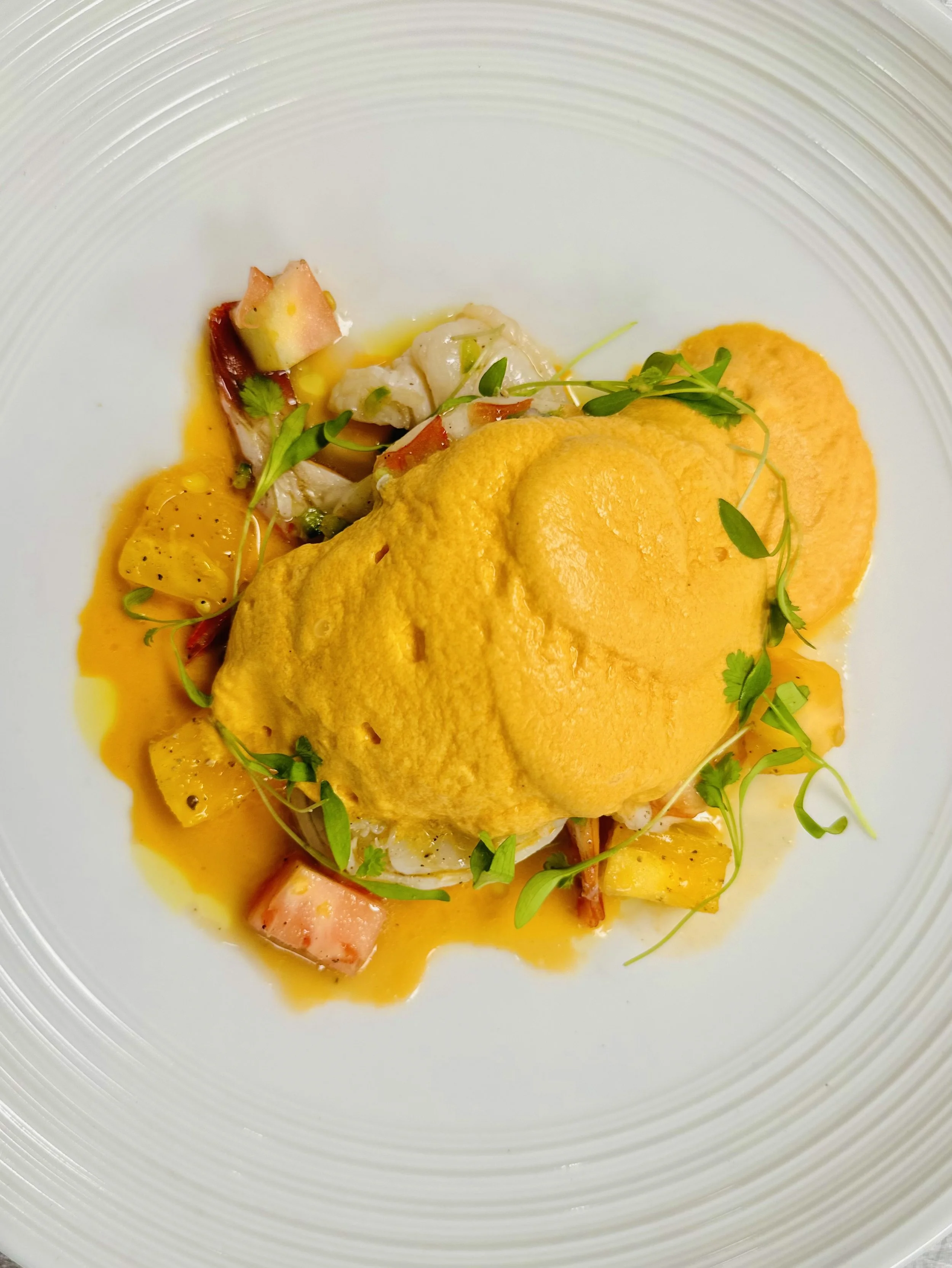 A plated dish featuring a dollop of bright yellow sauce, accompanied by pieces of seafood and garnished with microgreens and small vegetable chunks.