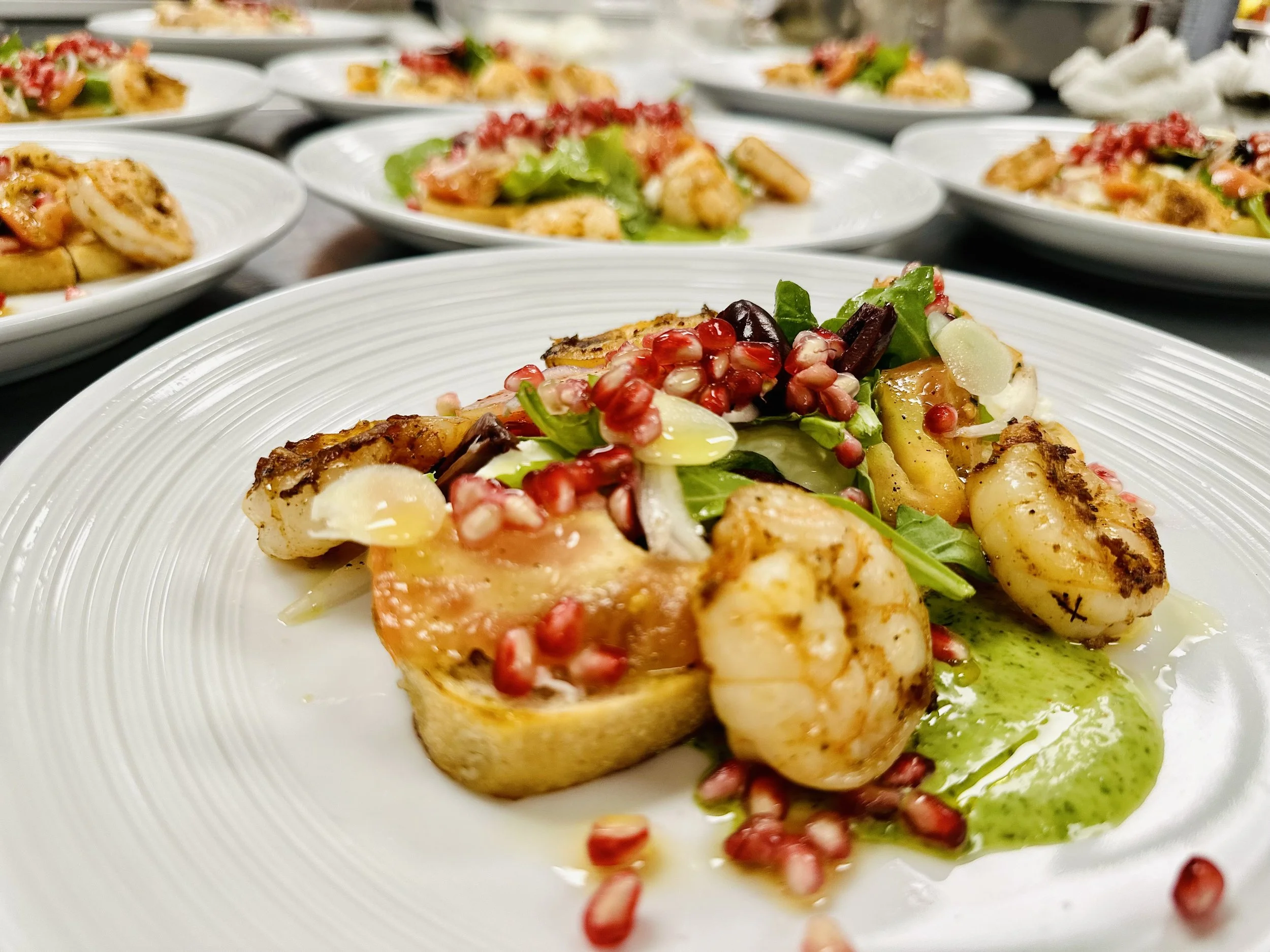 Close-up of a plate with a gourmet dish featuring scallops, garnished with pomegranate seeds, greens, and a green sauce, with multiple similar dishes blurred in the background.