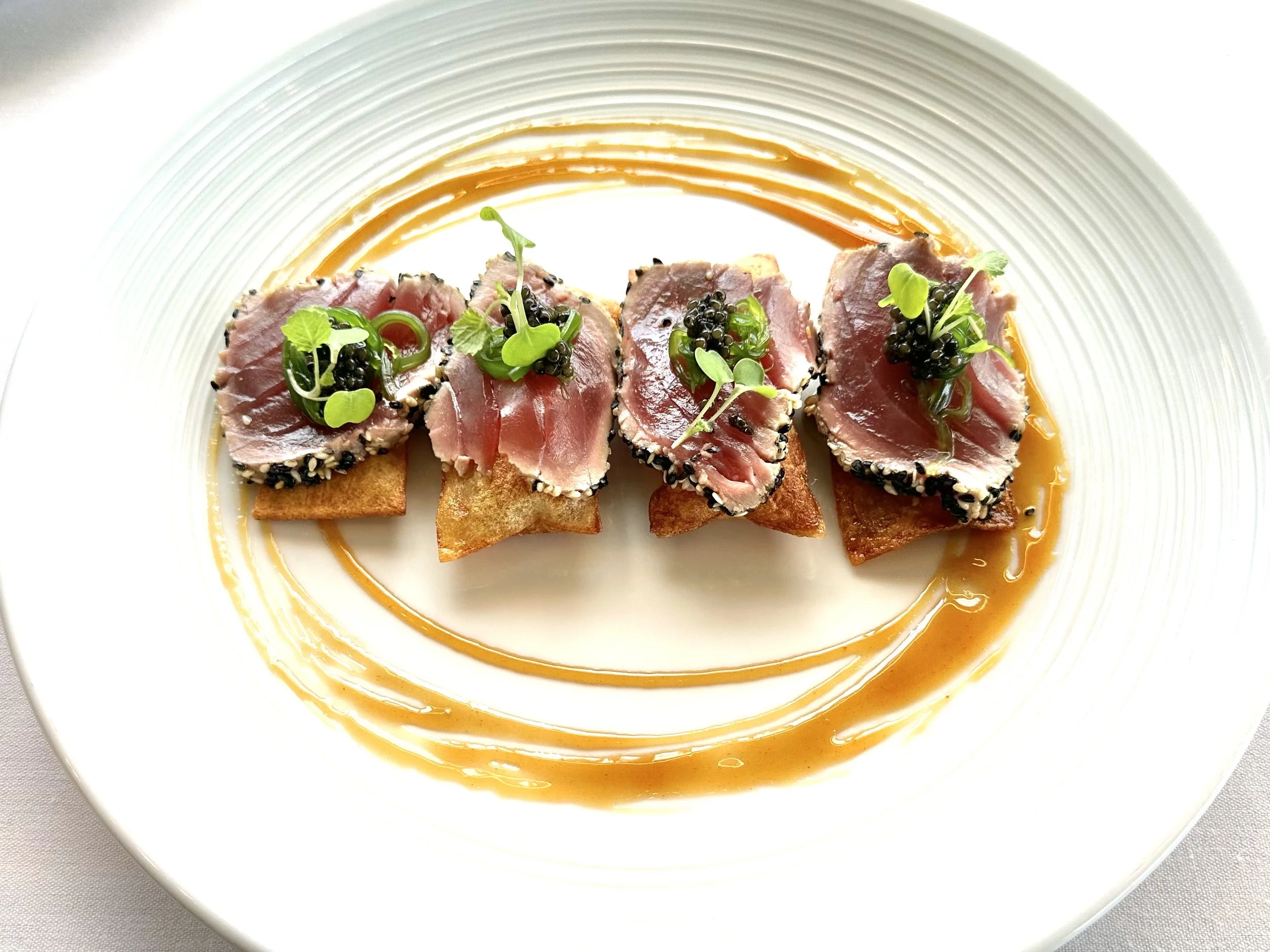 Seared tuna slices garnished with microgreens and caviar, served on toasted bread with a drizzle of orange sauce on a white plate.