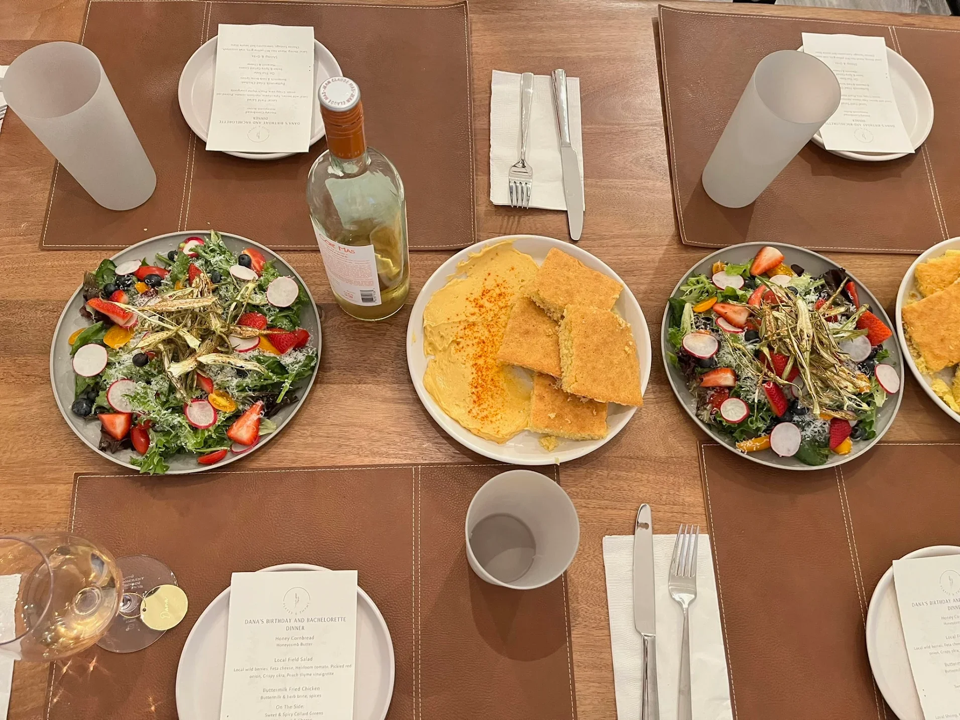 A table set for a meal with two large salads, a plate of breaded and fried food, a bowl of hummus garnished with paprika, and a bottle of wine, with cutlery, napkins, and empty glasses on place mats.