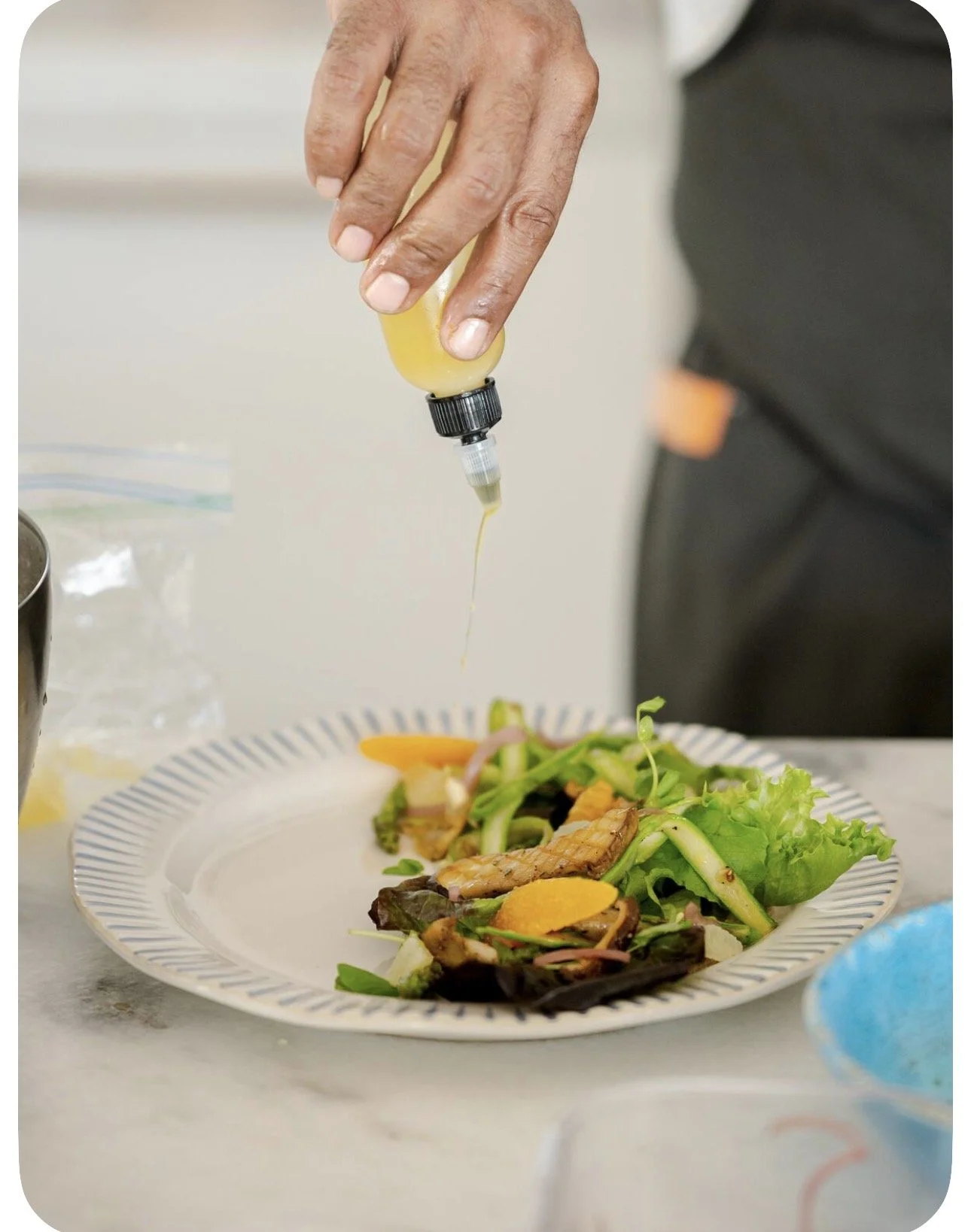 A person drizzling dressing or oil onto a plate of salad containing assorted vegetables including greens, carrots, and possibly grilled chicken.