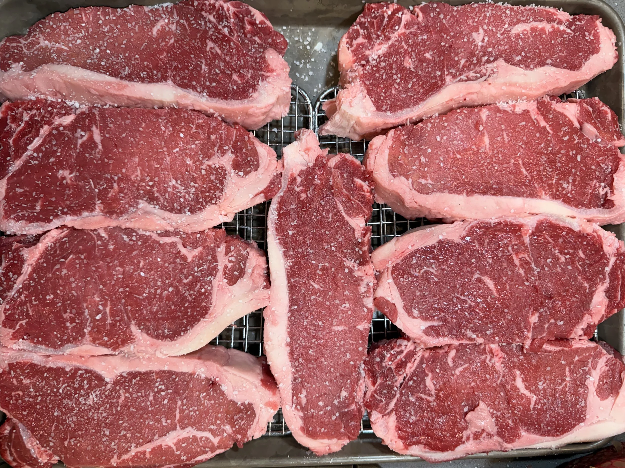 Four raw, seasoned beef steaks on a wire rack inside a metal tray.