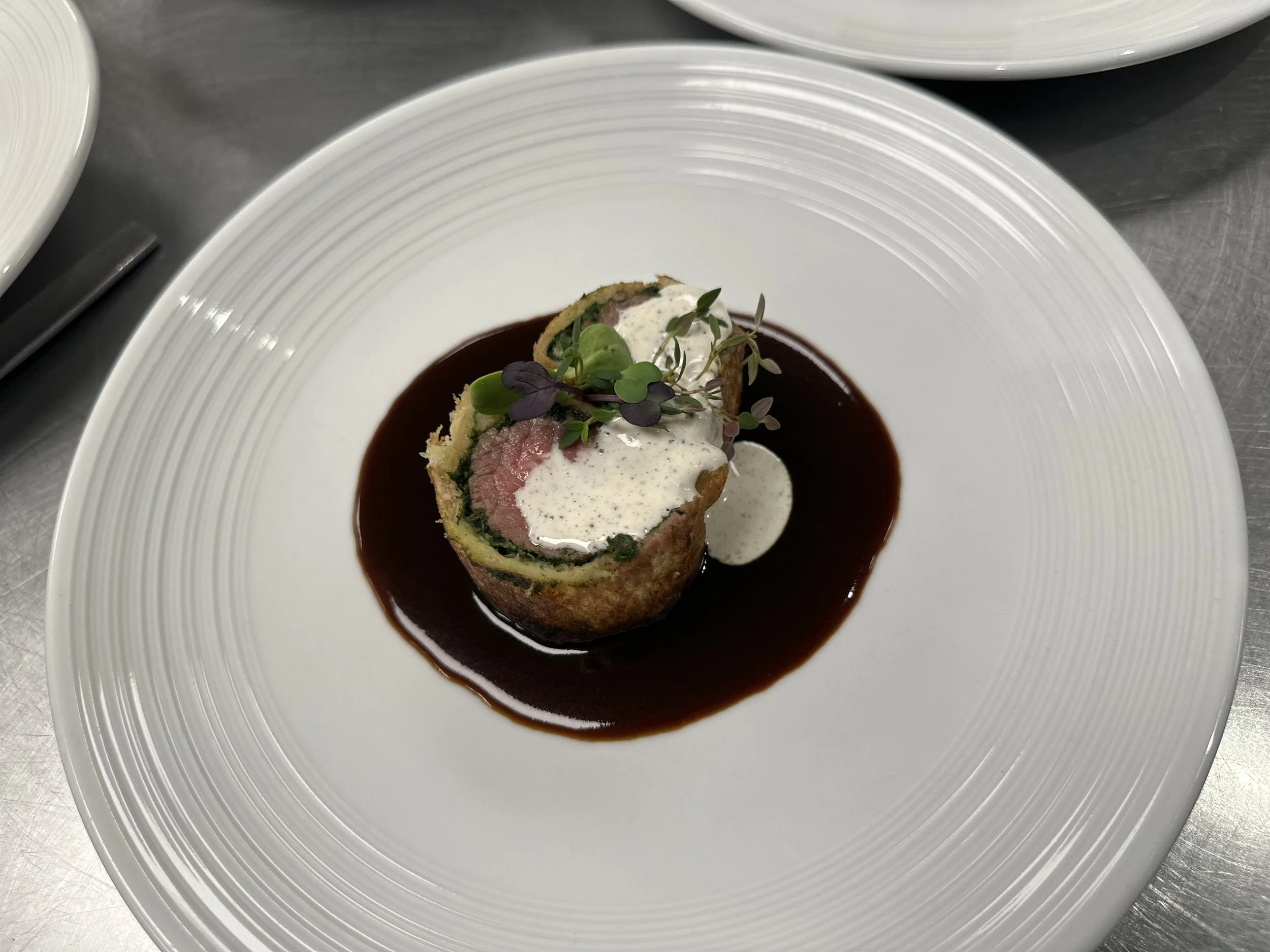 A plated gourmet dish consisting of a rolled meat pastry with sauce and microgreens, served in a white, textured bowl with a dark sauce at the base.