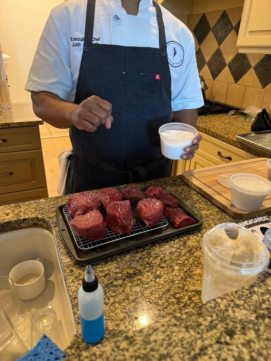 A chef in a white uniform and black apron standing behind a kitchen counter, holding a container of seasoning or white substance, with raw beef steaks on a tray in front. The kitchen has countertops, cabinets, and various cooking supplies.