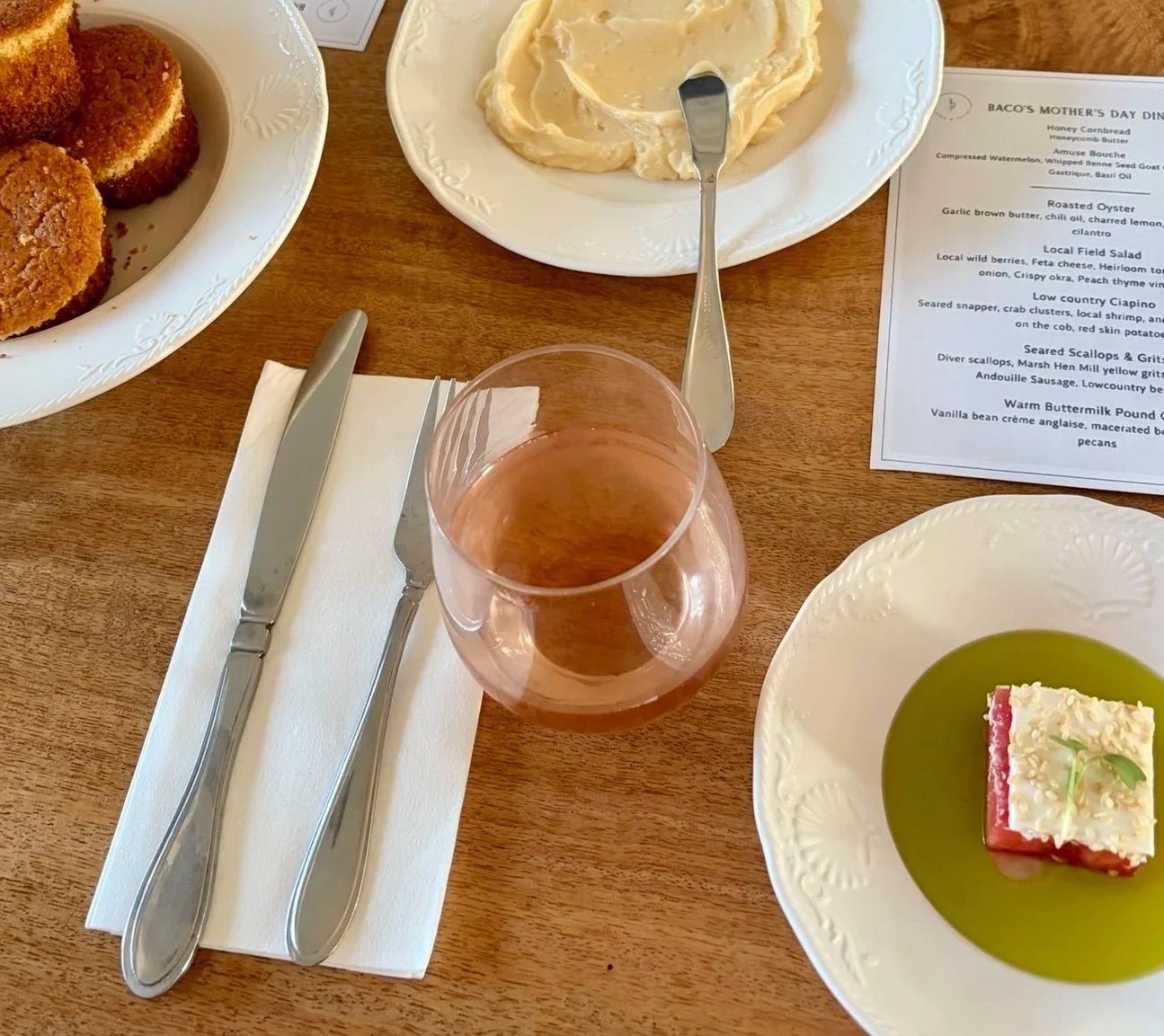 A table setting with a glass of rosé wine, a white napkin, and silverware. Alongside are plates with food, including fried croquettes, mashed potatoes with a serving spoon, and a square dessert in green sauce. A printed menu is visible on the table.